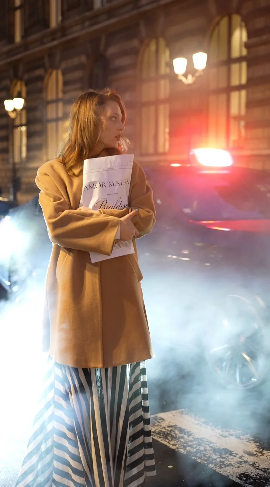A woman with shoulder-length hair in a beige coat holding a script, standing on a city street at night next to a police car with flashing lights and fog.