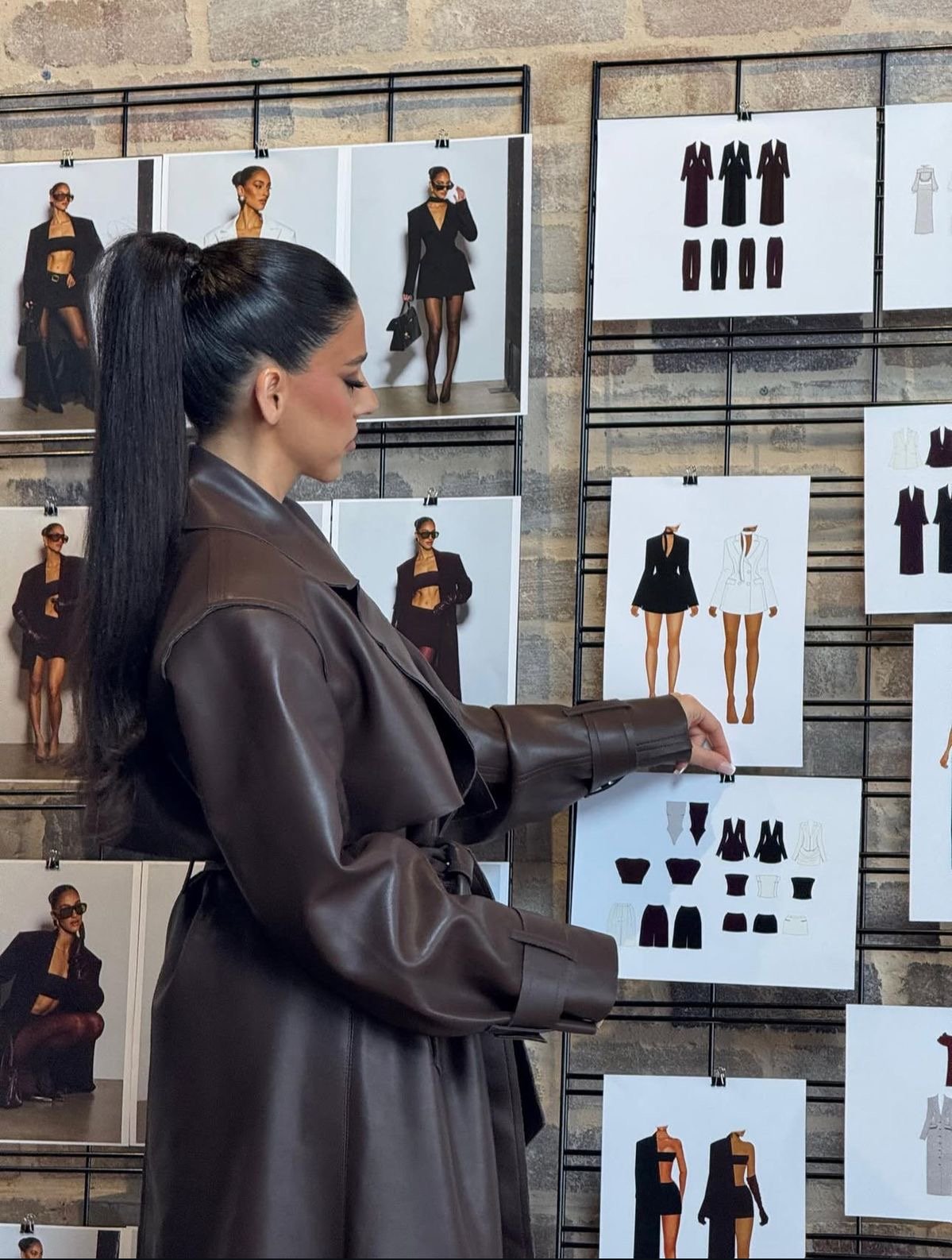 A woman wearing a black blazer and black skirt standing in front of a white wall with decorative molding and a white mantel with a white vase with black flowers.
