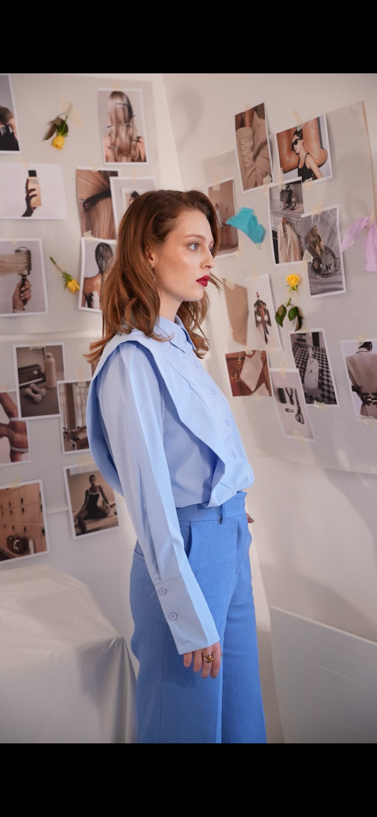 A woman with red hair wearing a light blue blouse and matching blue pants, standing in front of a wall decorated with various photos and flowers.