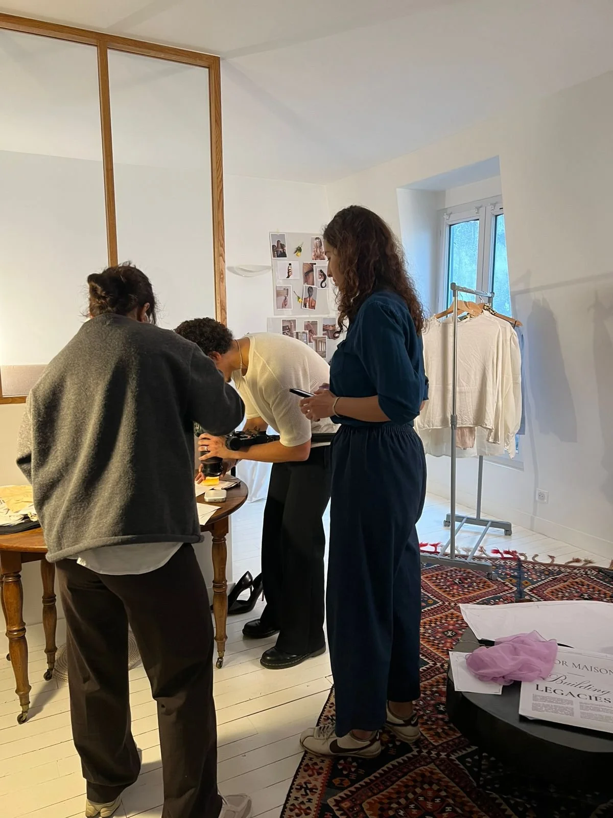 Three people standing around a table in a well-lit room. One person is leaning over to examine items on the table, while another is standing and another is holding a phone. There is a clothing rack with white garments and a window in the background. The room has a patterned rug and a wall decorated with photos.