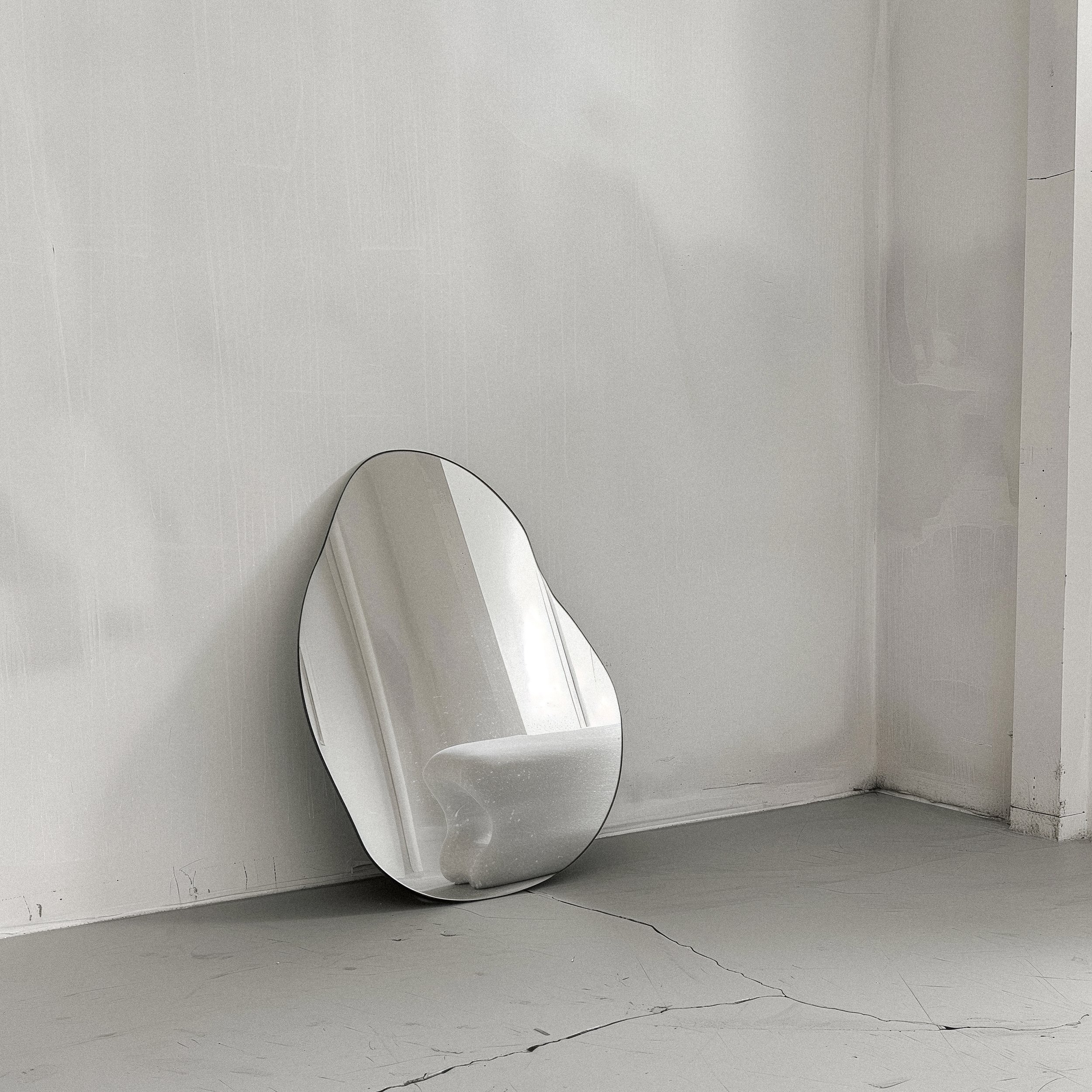 A modern, irregularly shaped mirror leaning against a plain, light-colored wall in a minimalistic room with cracked flooring.