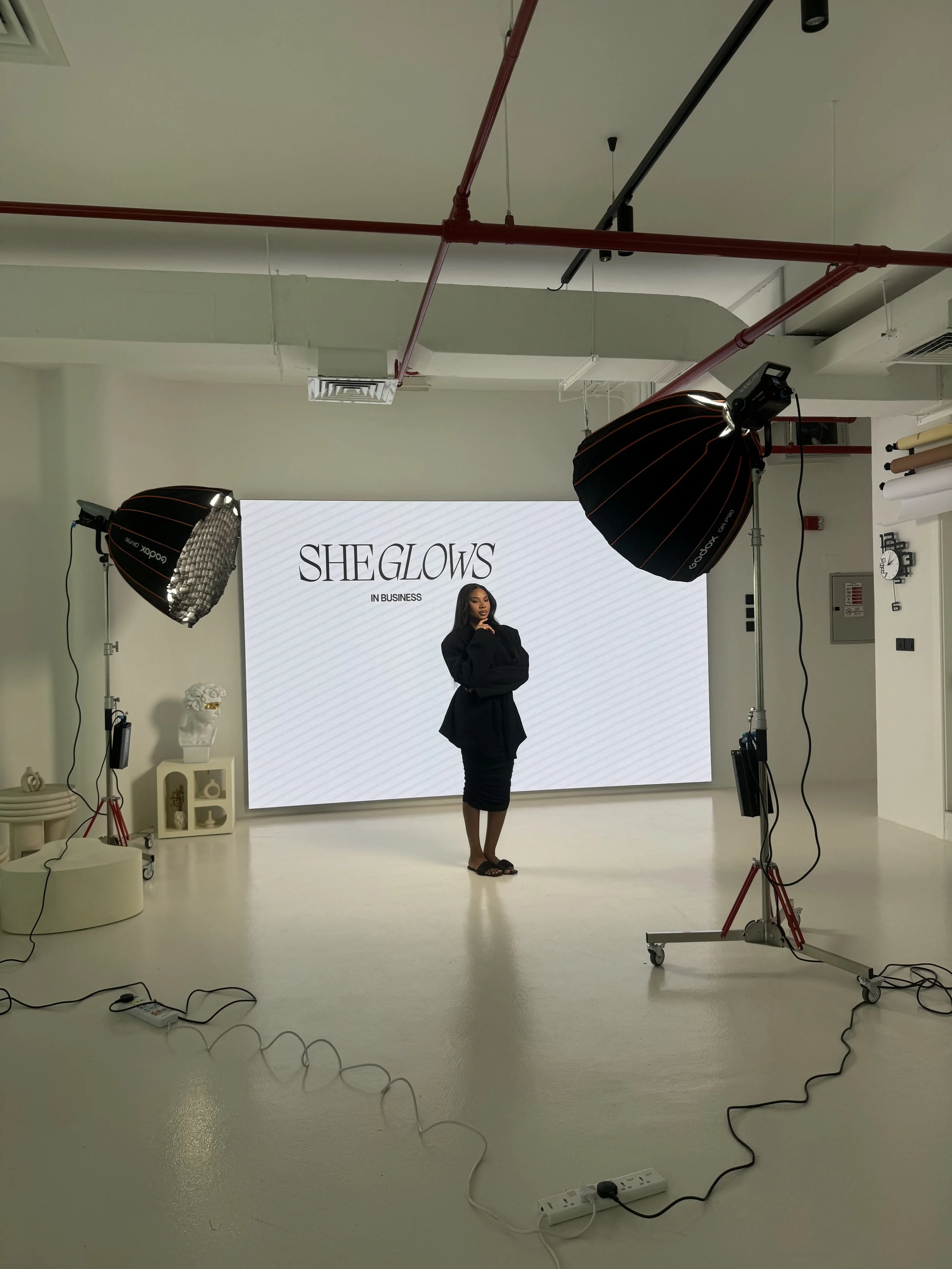 People gathered around a table in a bright room with white walls, a window, and a piece of clothing on a rack, possibly preparing for a photo shoot or interview.