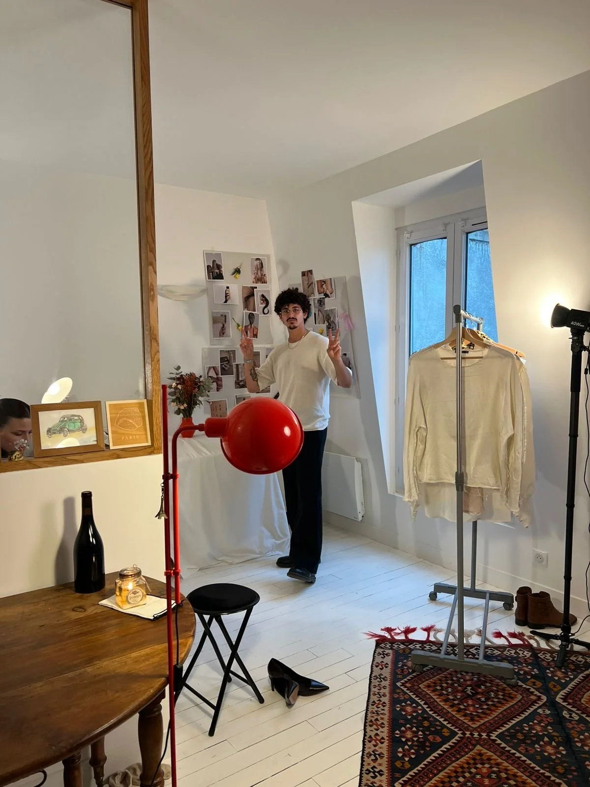 A man with curly hair and glasses making peace signs in a room with a white wall decorated with photos, a red desk lamp, a wooden dining table with a black vase, candles, and pictures, a rack with cream-colored clothing, a window, a colorful rug, and