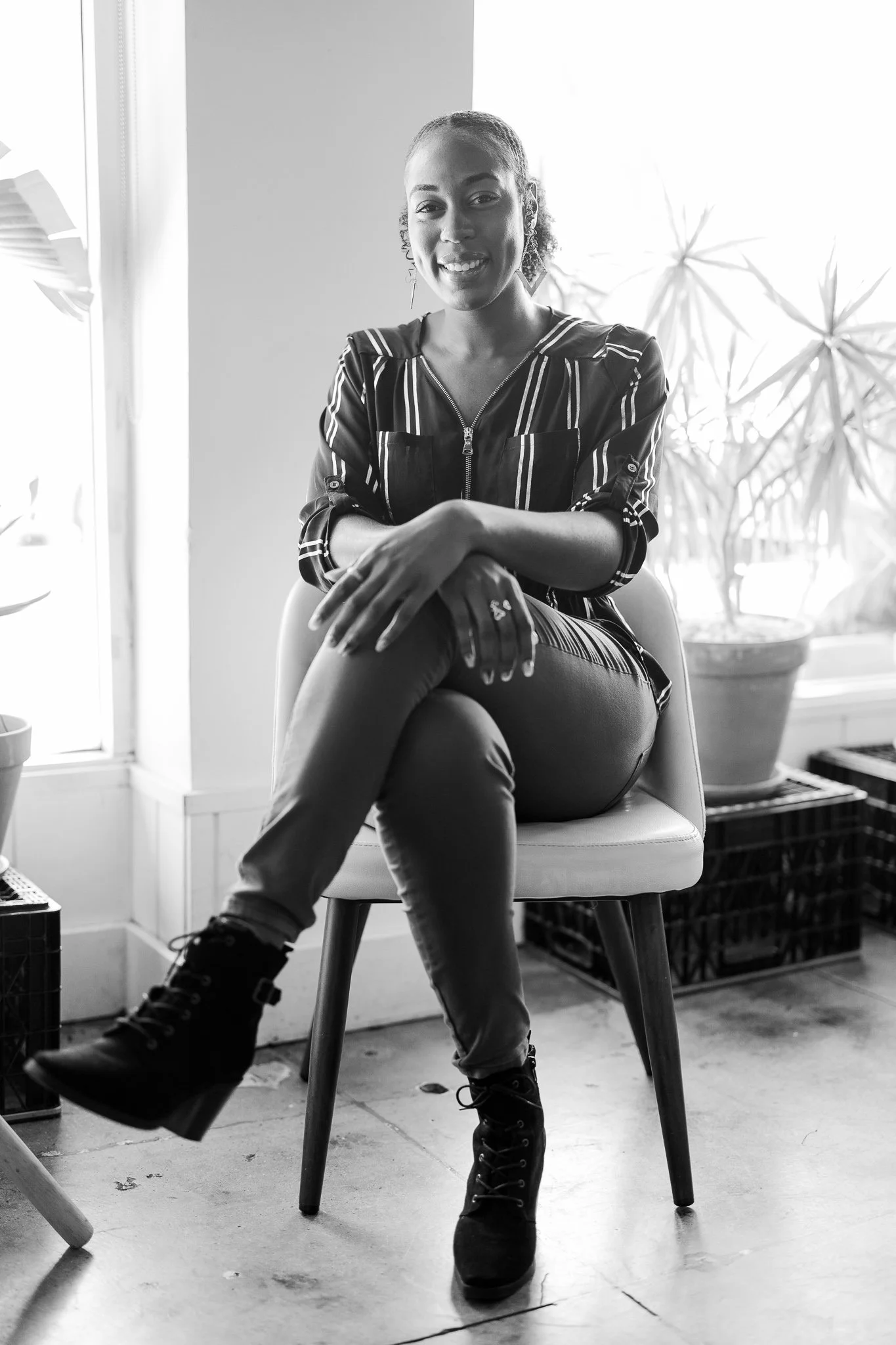 A woman sitting on a chair indoors, smiling at the camera, with potted plants and crates in the background.