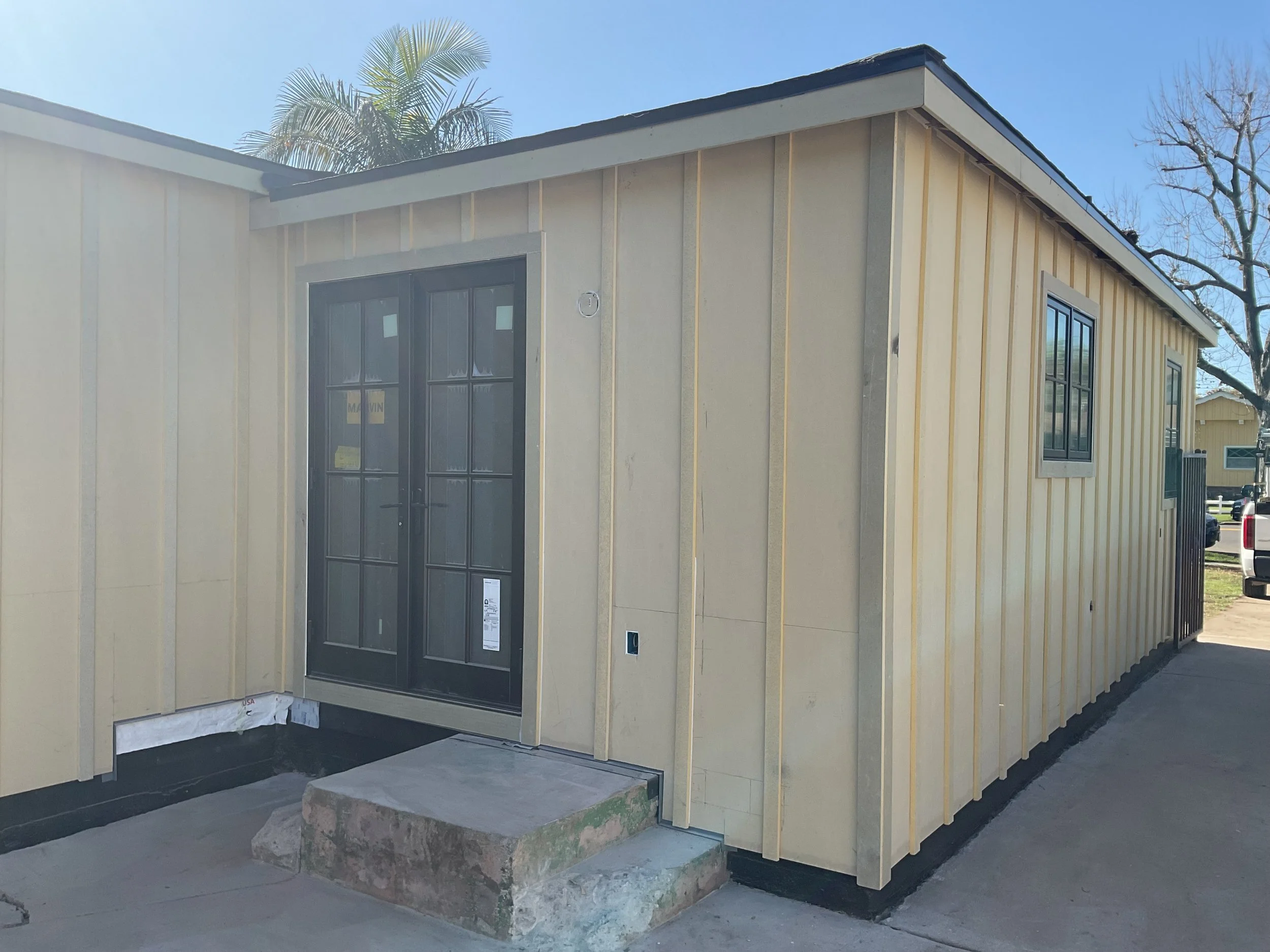Newly constructed beige house with black trim, built with vertical siding panels, featuring two black-framed windows and a black front door, situated on a concrete foundation with a large rock in front.