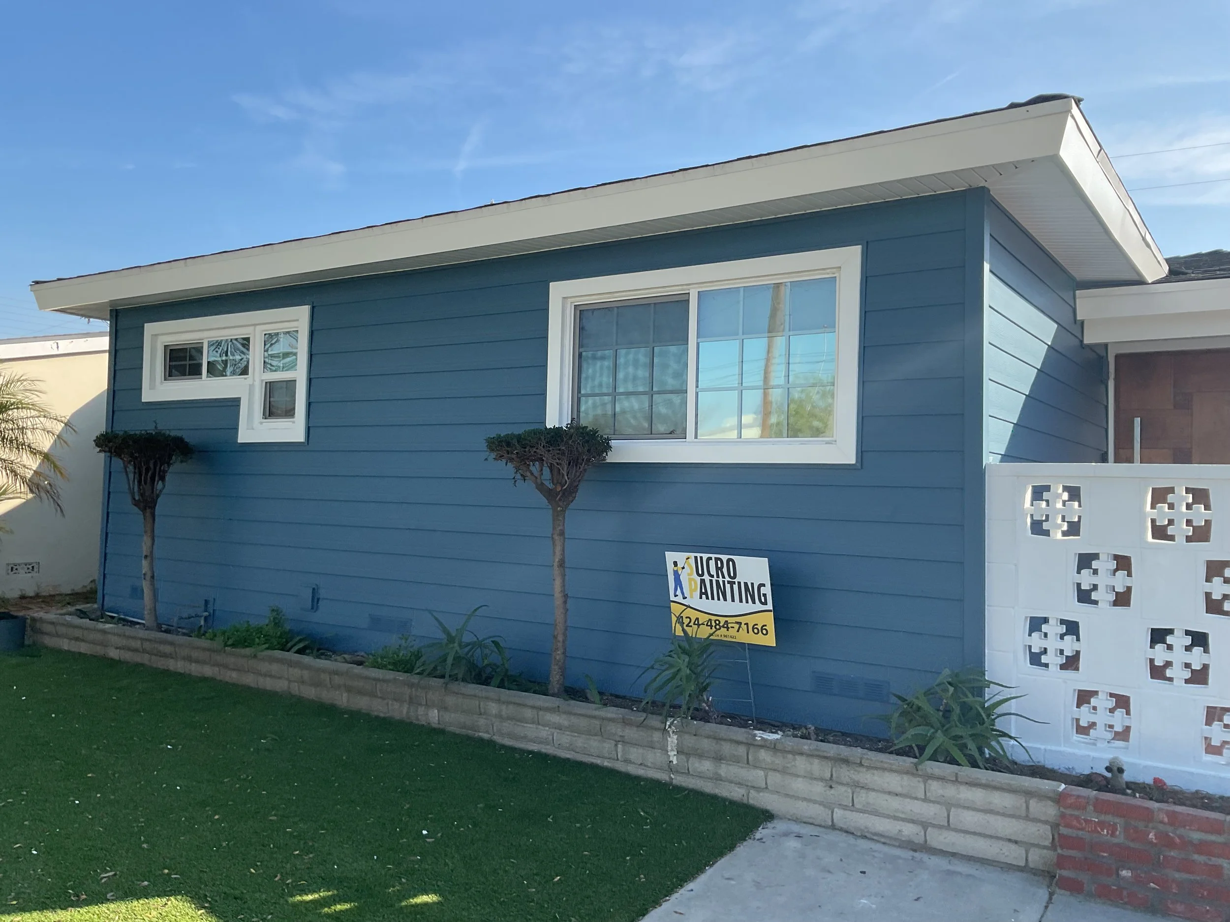 A blue house with large windows, a small window, and a white roof. There are two small trees in front of the house and a garden bed with plants. A yellow sign advertising UCRO painting is in the garden with a phone number.