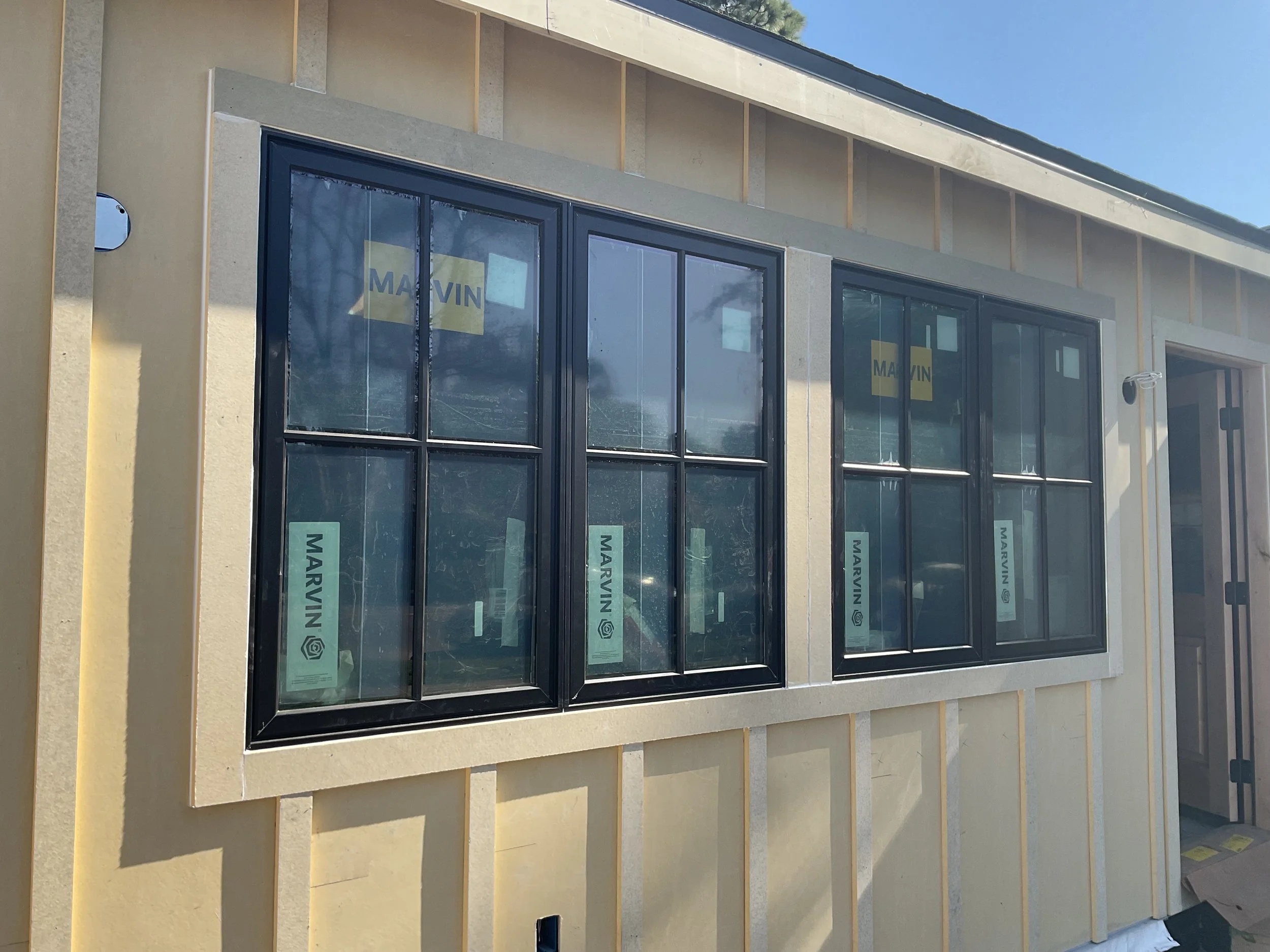 Exterior of a building under construction with large black-framed windows and yellow insulation panels. Some windows still have the brand name 'MARVIN' visible on the protective stickers.