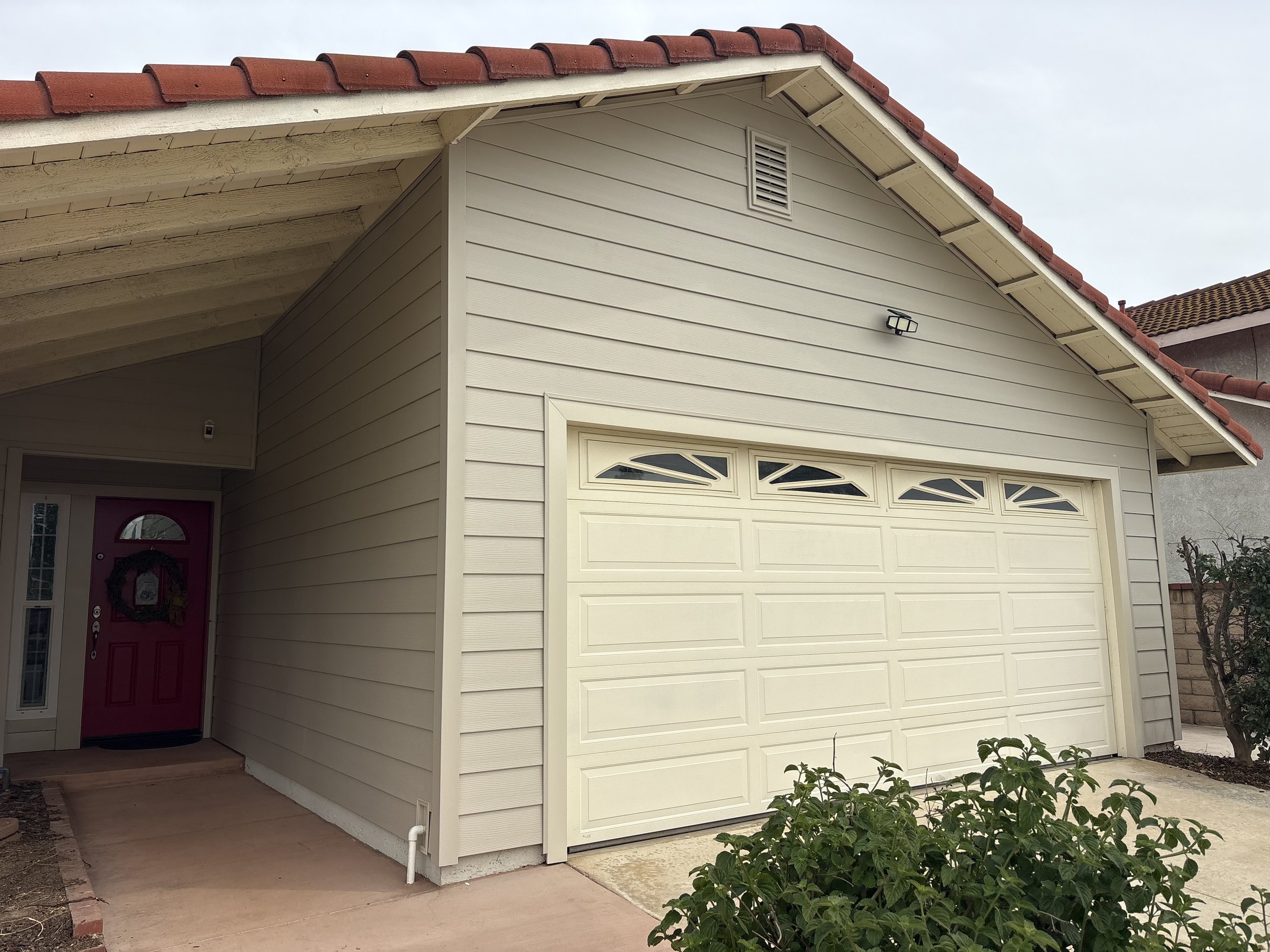 Exterior view of a house with a beige garage door, cream siding, and a red front door. The house has a sloped roof with red tiles, and a small vent near the roof's peak. There is a black outdoor light mounted on the wall above the garage door. The driveway is concrete, and there are some plants and a small tree in the garden area.