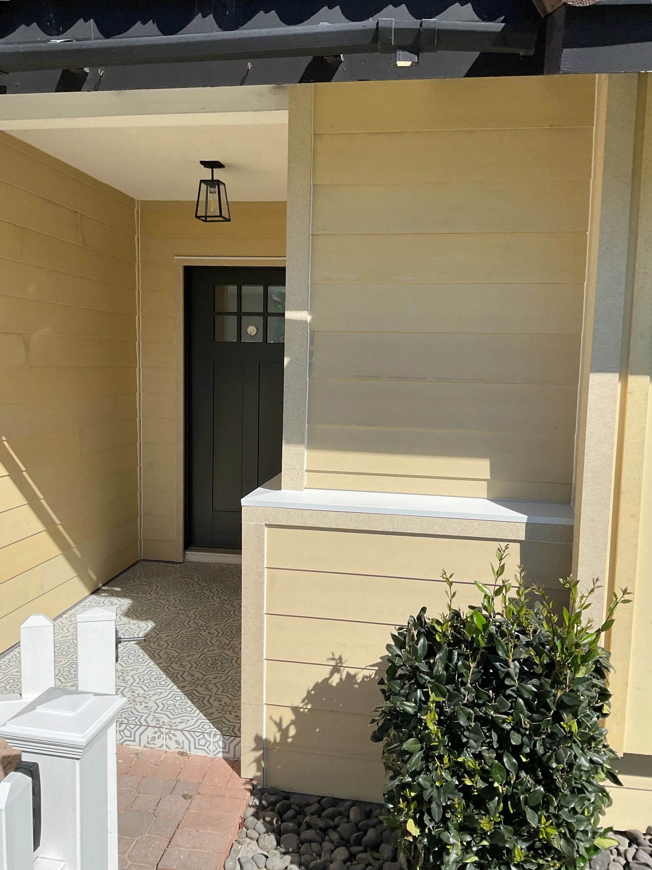 Front porch area of a house with a black door, light yellow siding, and a small covered porch with hanging light fixture. There is a short wall with a planter box, a bush, and a decorative tiled walkway.