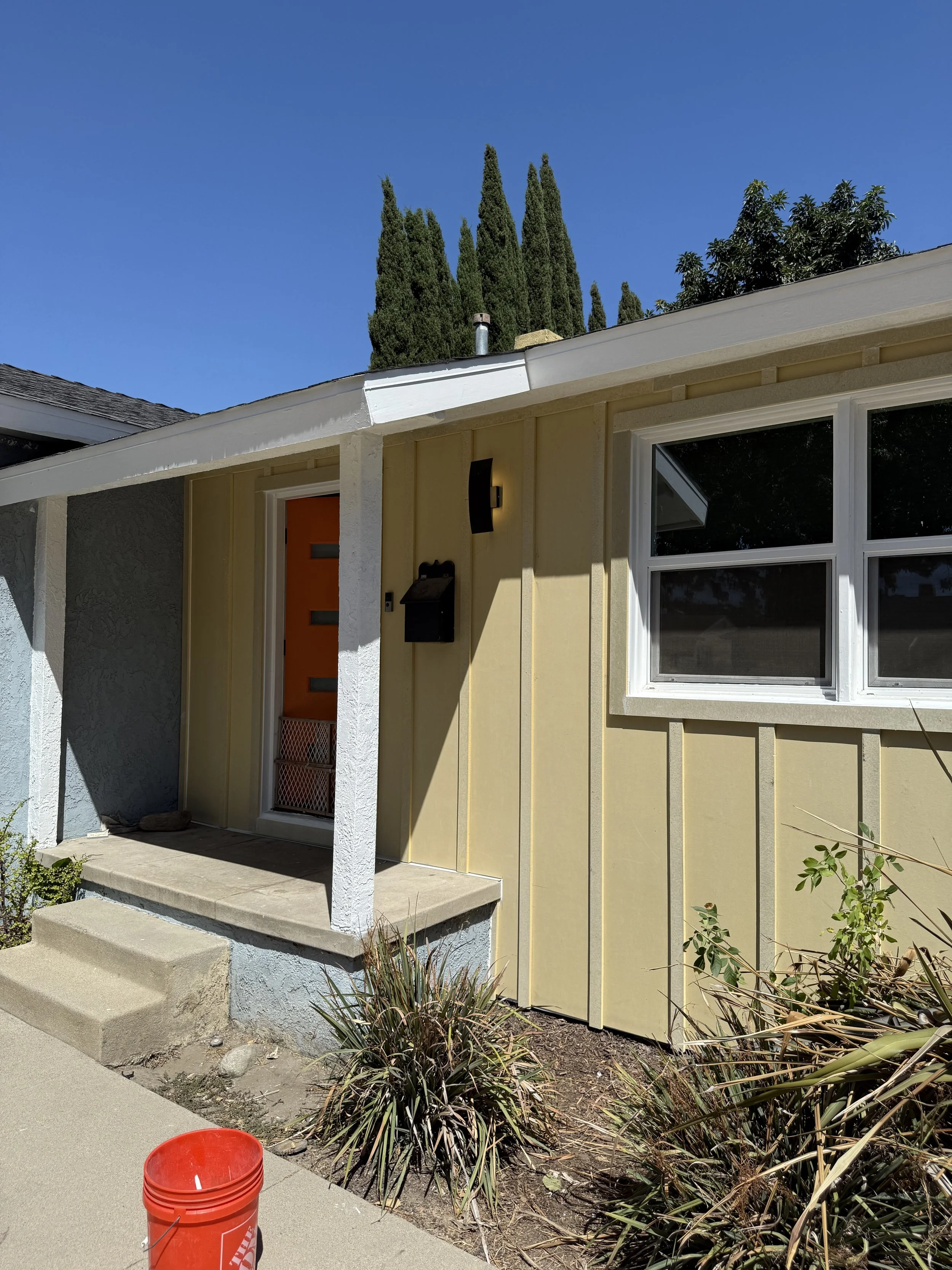 A house with a yellow exterior and white trim, featuring a small porch with steps leading up to the front door. There are tall, dark green cypress trees in the background under a clear blue sky. In the foreground, there are some plants and a red bucket on the sidewalk.