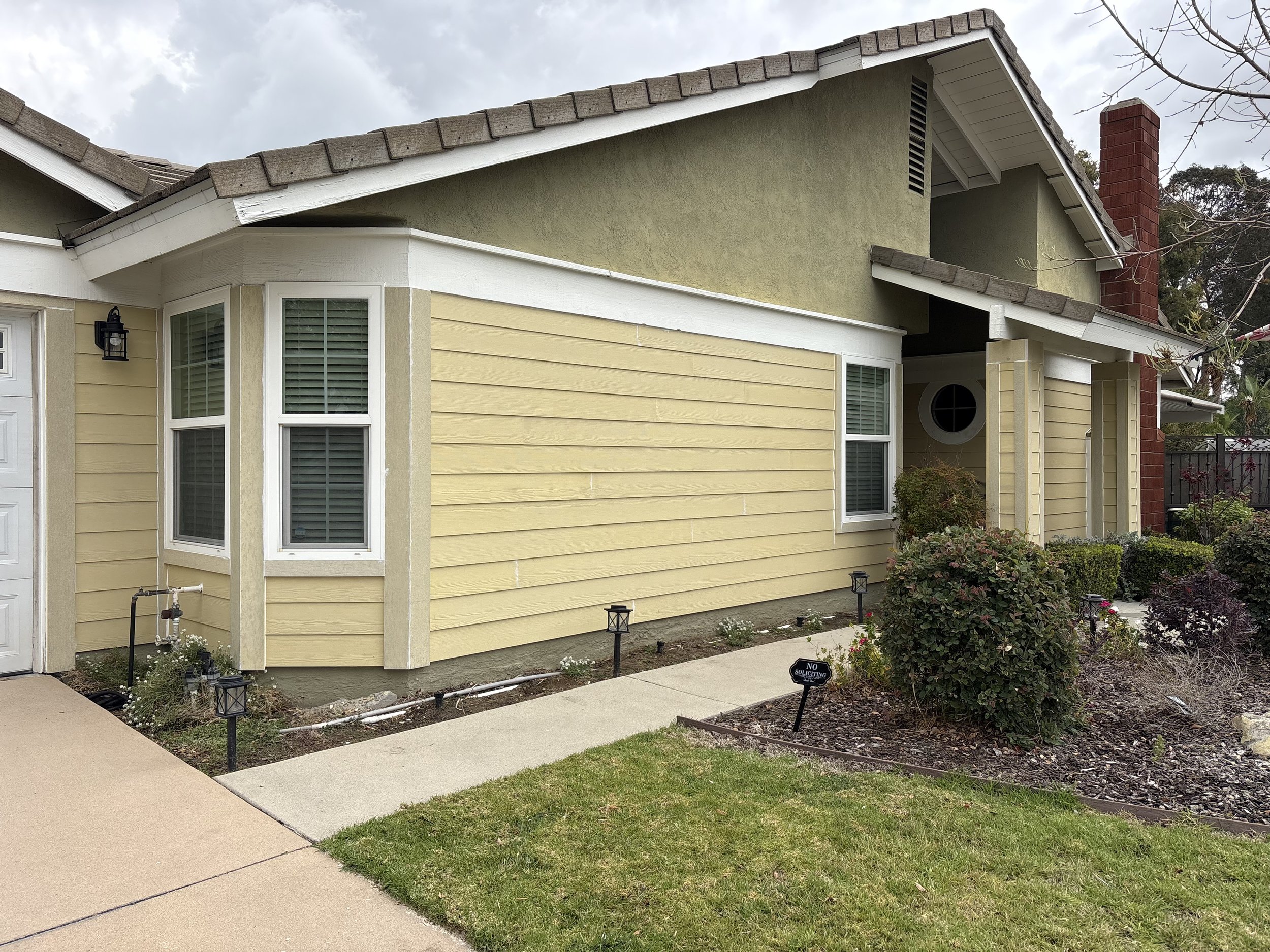 Front yard of a house with yellow siding and green stucco upper section, white window trim, a small garden, pathway, and a black 'No Soliciting' sign.