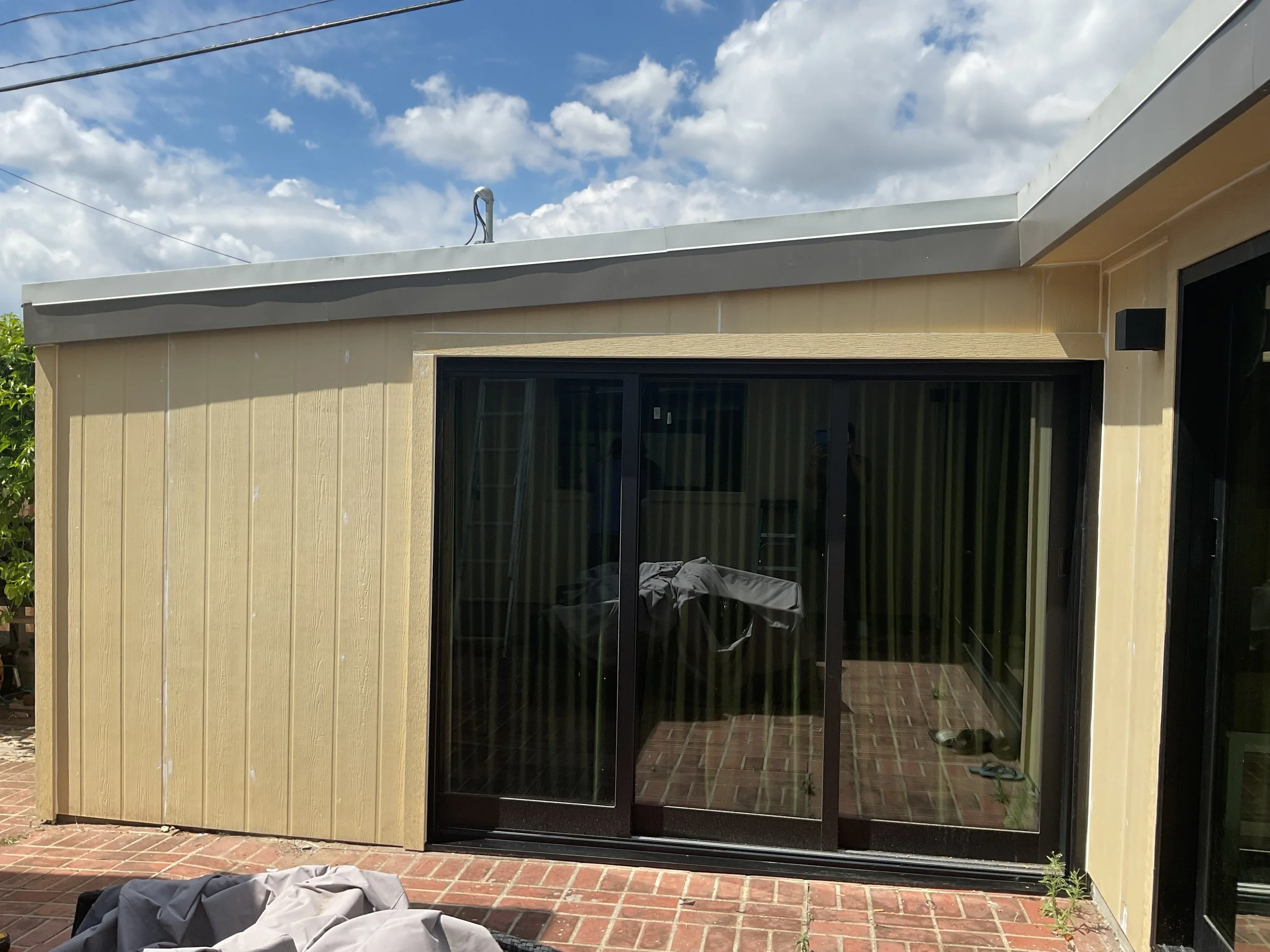 The exterior of a house with beige siding, a black sliding glass door, and a covered patio area with brick flooring. A gray fabric-covered object and some shoes are on the ground in front, with a blue sky and clouds overhead.