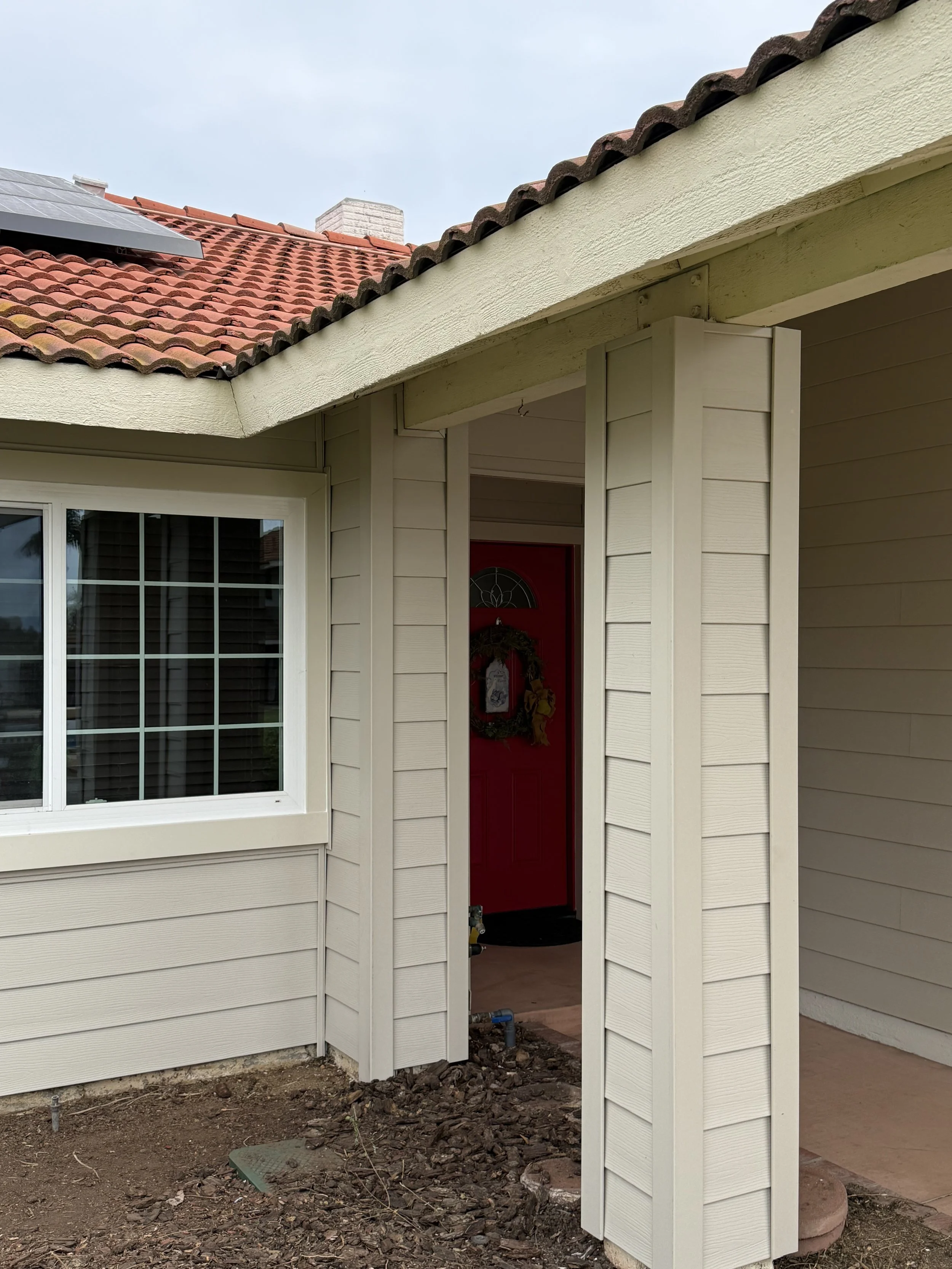 The front porch area of a house with beige siding, a red door with a wreath, a large window, and a roof with terracotta tiles. There is some construction or landscaping work happening in the foreground.