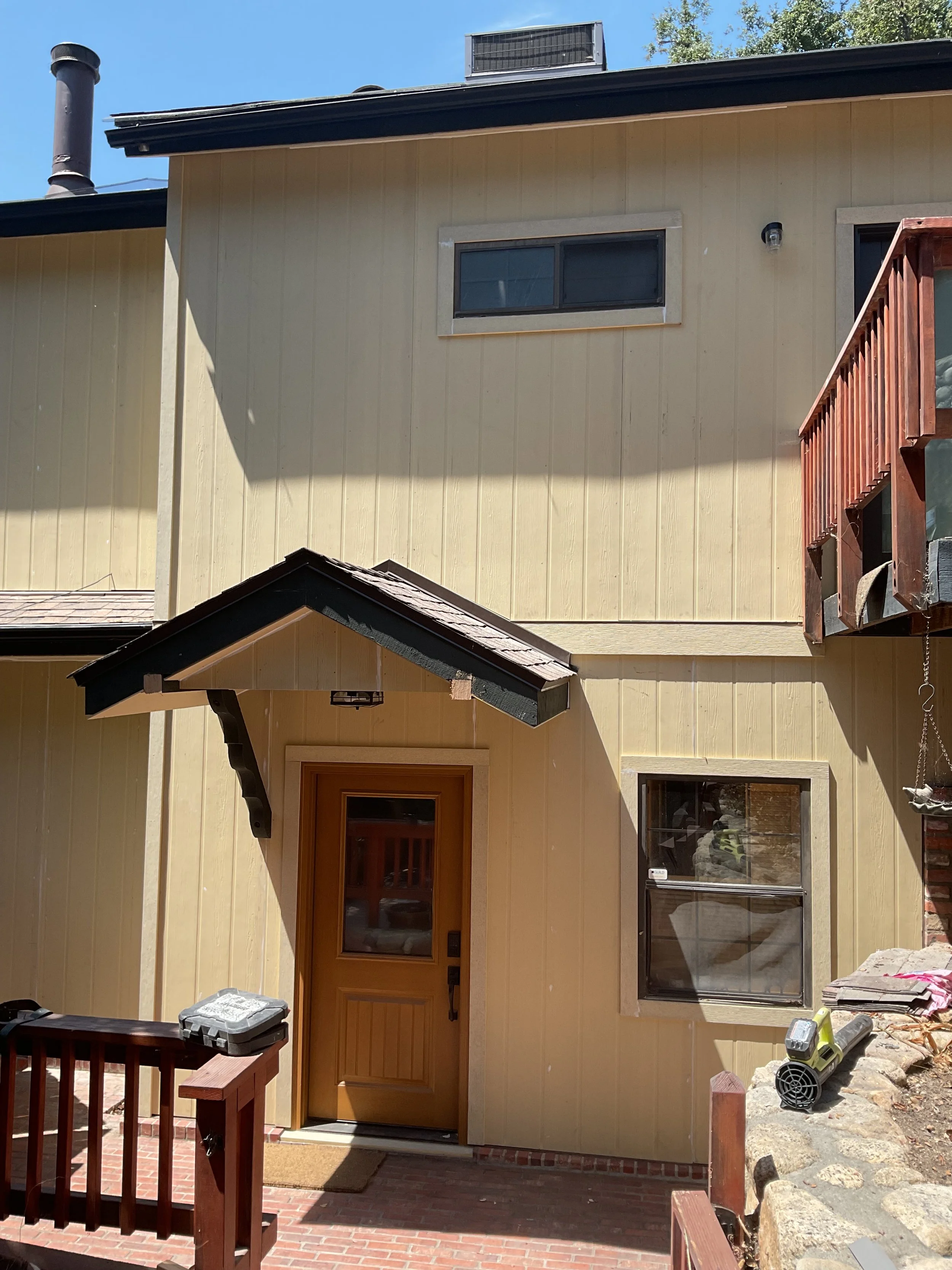 Close-up view of the front entryway of a house painted in yellow with a wooden door and two windows, with construction tools and materials visible on the porch and surrounding area.