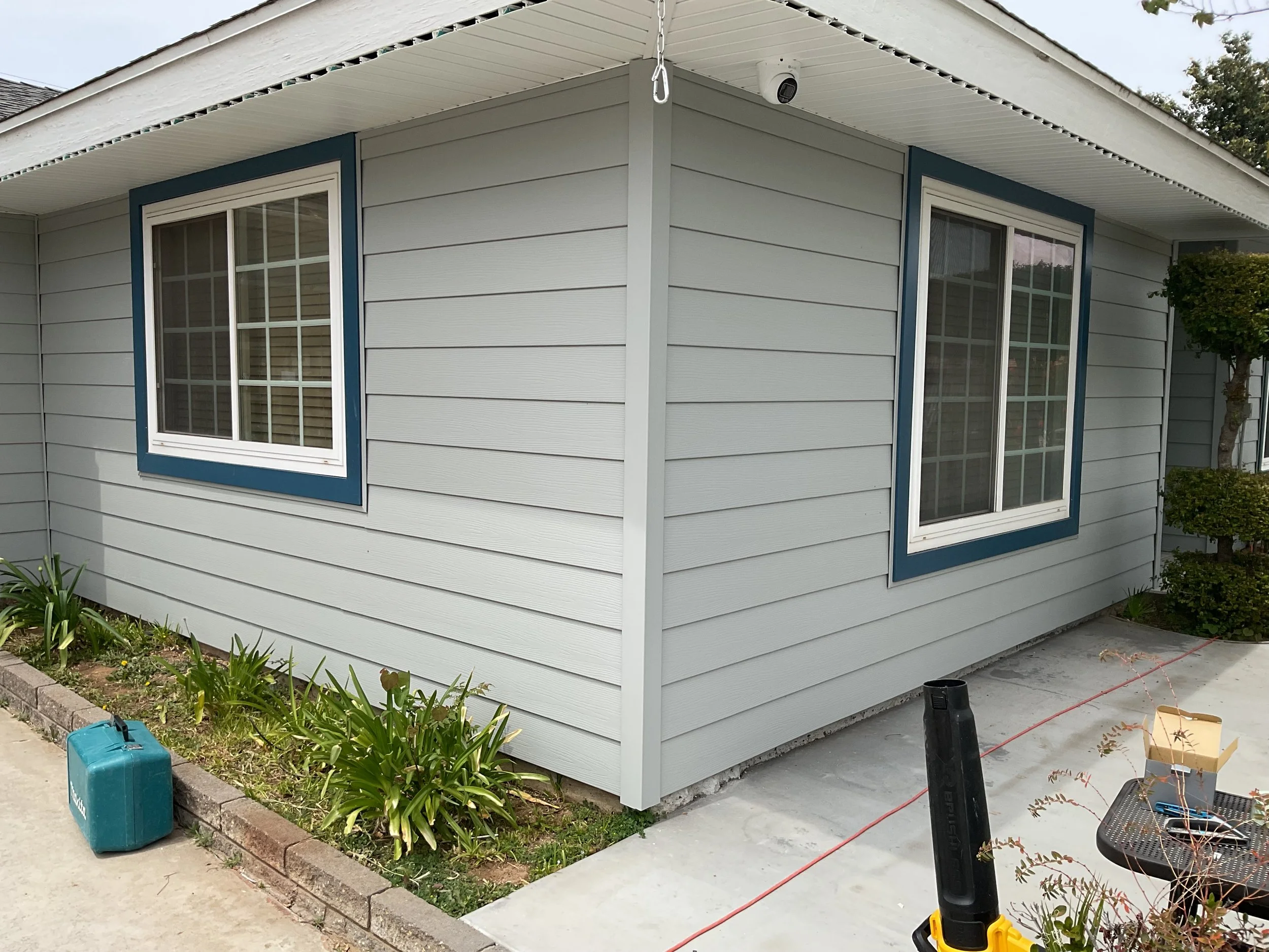 Close-up of a house corner with grey siding and white trim, featuring two large windows with white frames, a security camera, and a chain hanging from the roof's edge, surrounded by a small garden with plants and gardening tools nearby.
