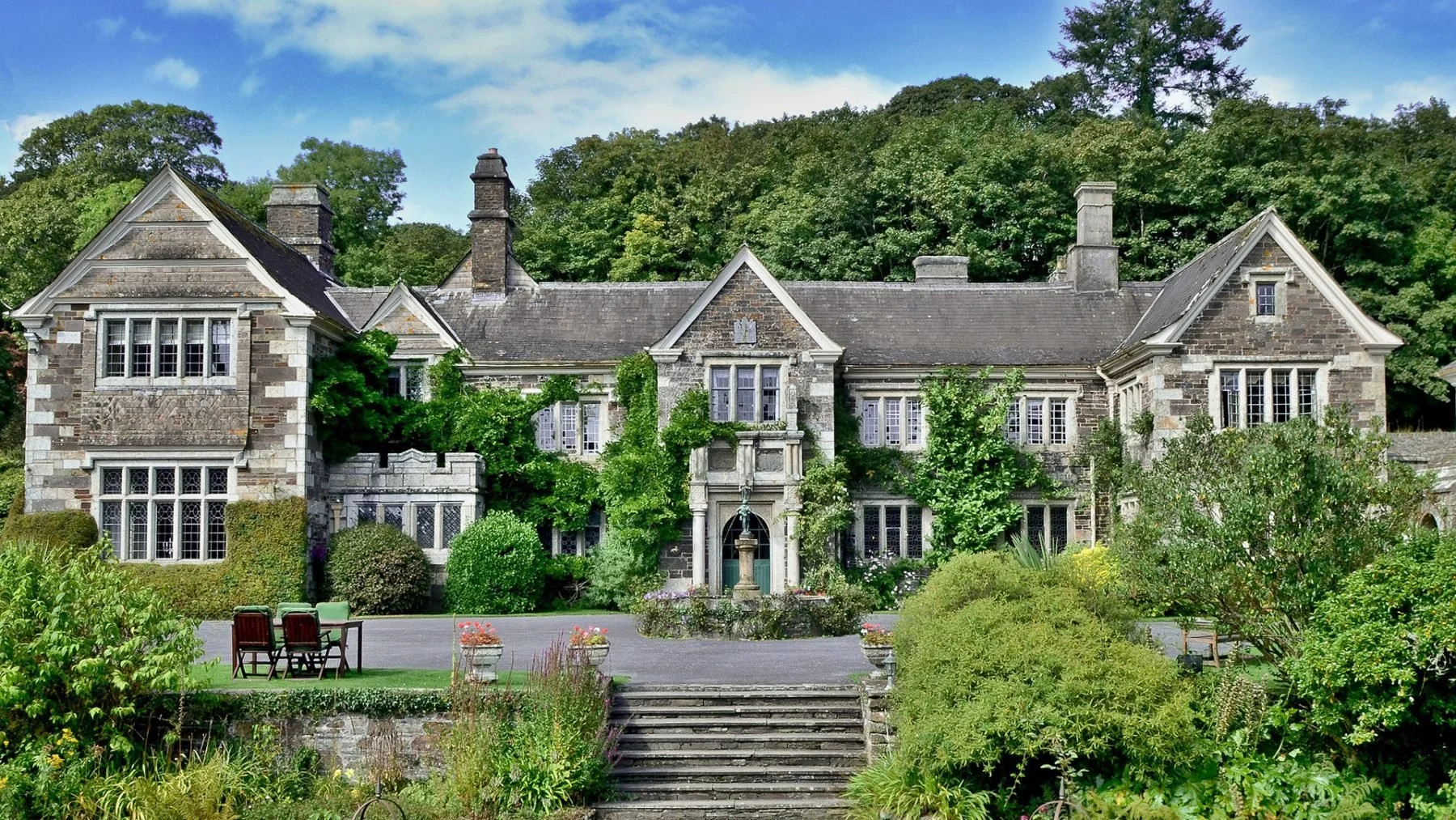 Large historic stone mansion with ivy, surrounded by lush greenery and garden, against a backdrop of trees.