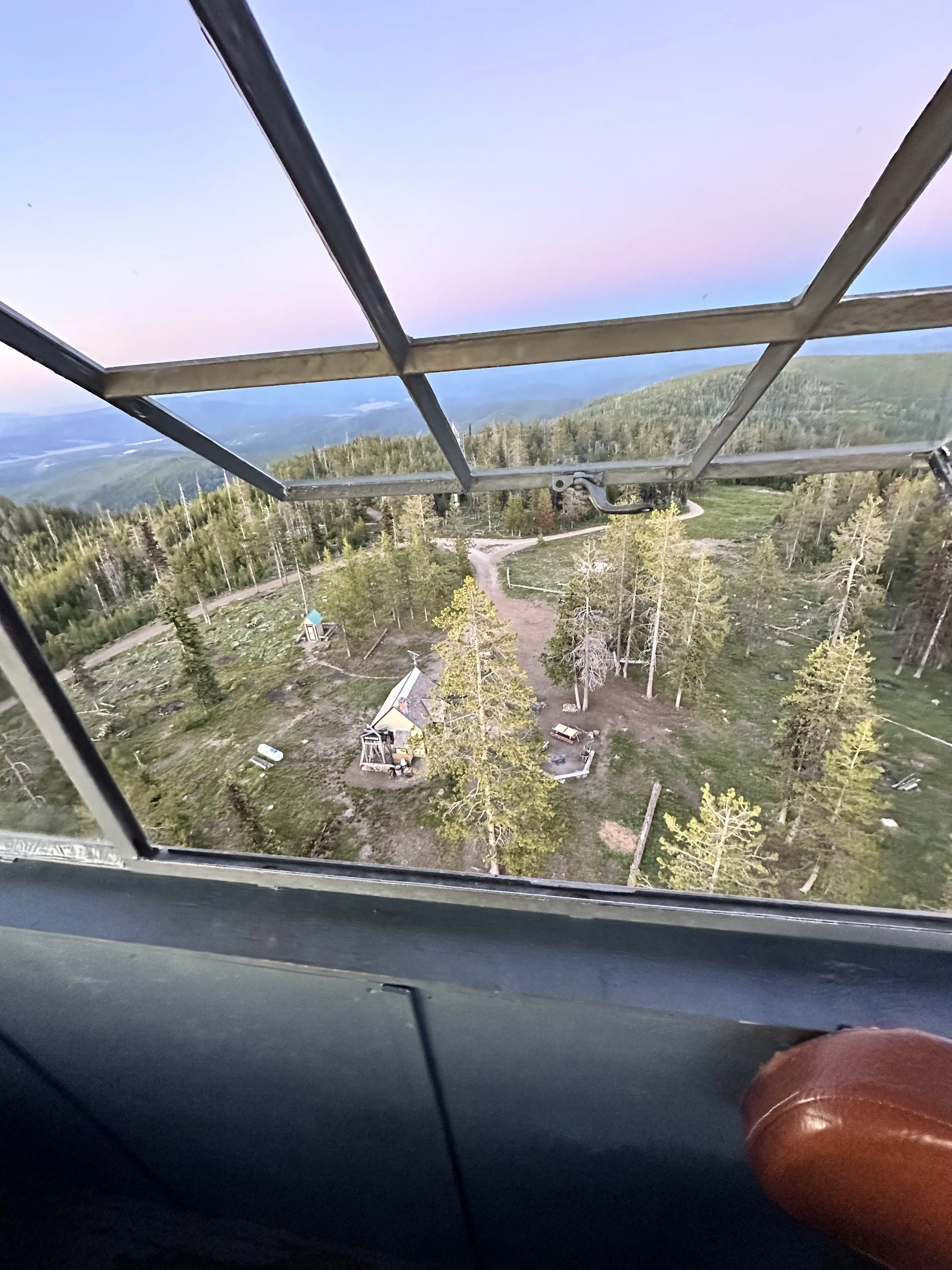 View from a lookout tower showing a forested landscape with trees, a small cabin-like structure, and winding dirt paths. The sky displays a gradient of colors from pink to blue, indicating early morning or late evening light.