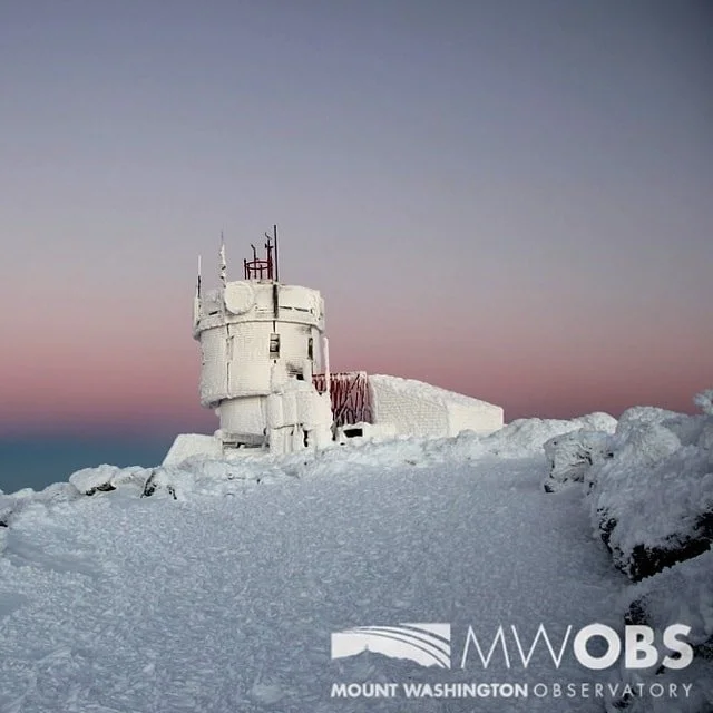 Mount Washington Observatory tower covered in snow with a pink and purple sky in the background.