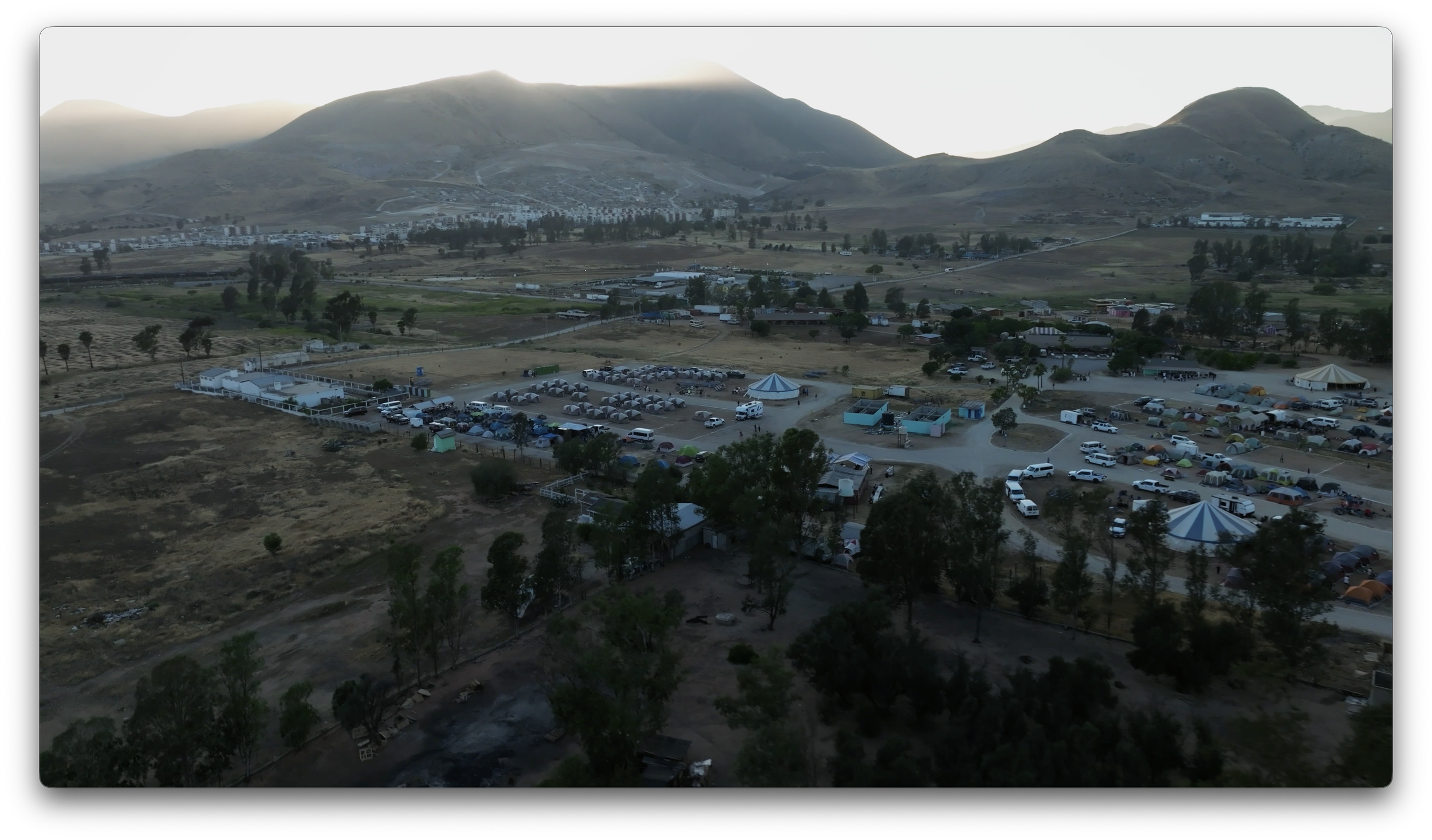 A panoramic view of a campground with tents, RVs, and parked vehicles, set against a backdrop of rolling hills and mountains.