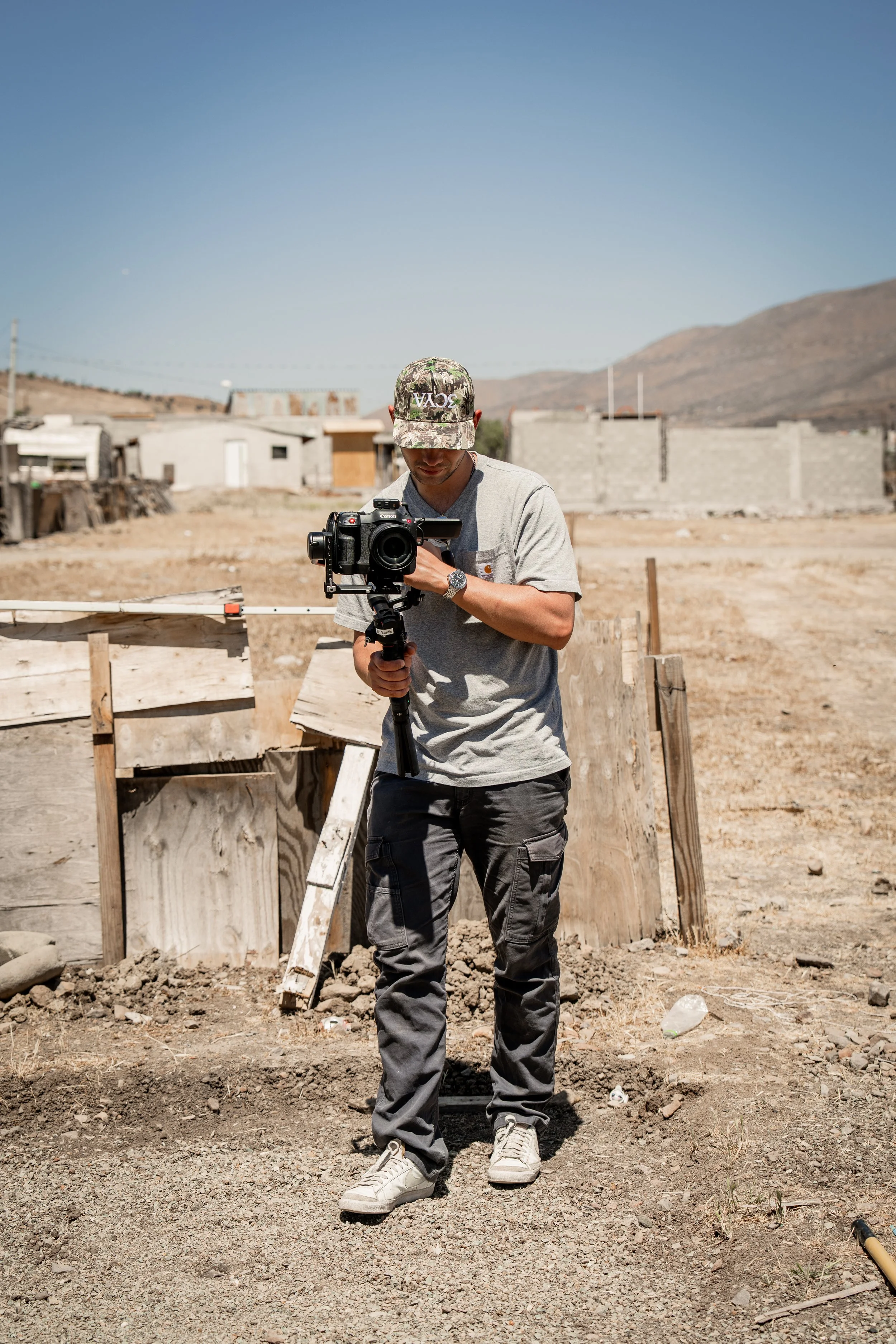 A man in a gray t-shirt, gray cargo pants, white sneakers, and a camouflage baseball cap operating a camera on a stabilizer in a dry, dusty outdoor construction site with makeshift wooden barriers and a rural background with hills and scattered buildings.