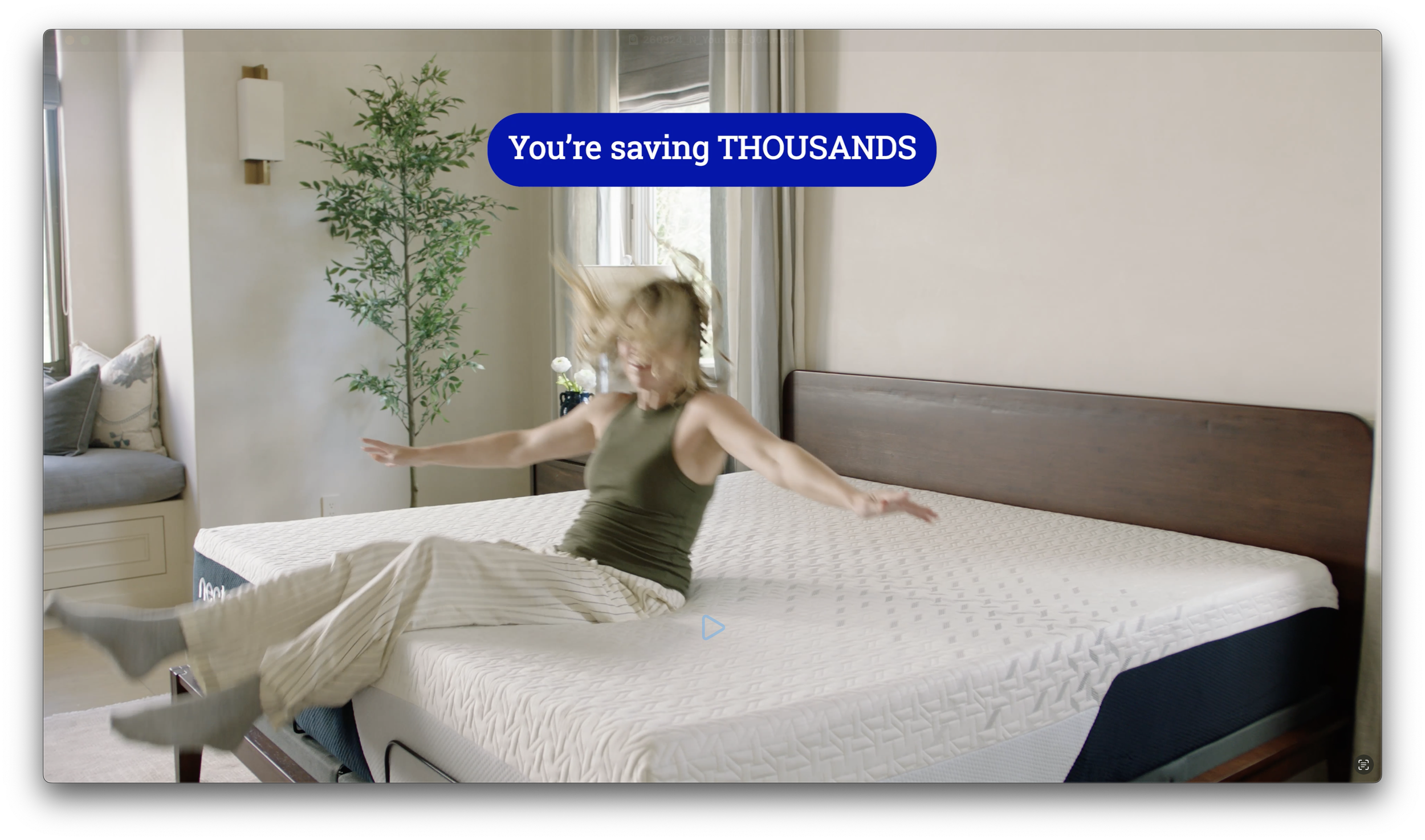 Person in gray tank top and light-colored pajama pants joyfully jumping on a bed in a bright bedroom with a large window, curtain, a potted plant, a wall sconce, and a bench with pillows.