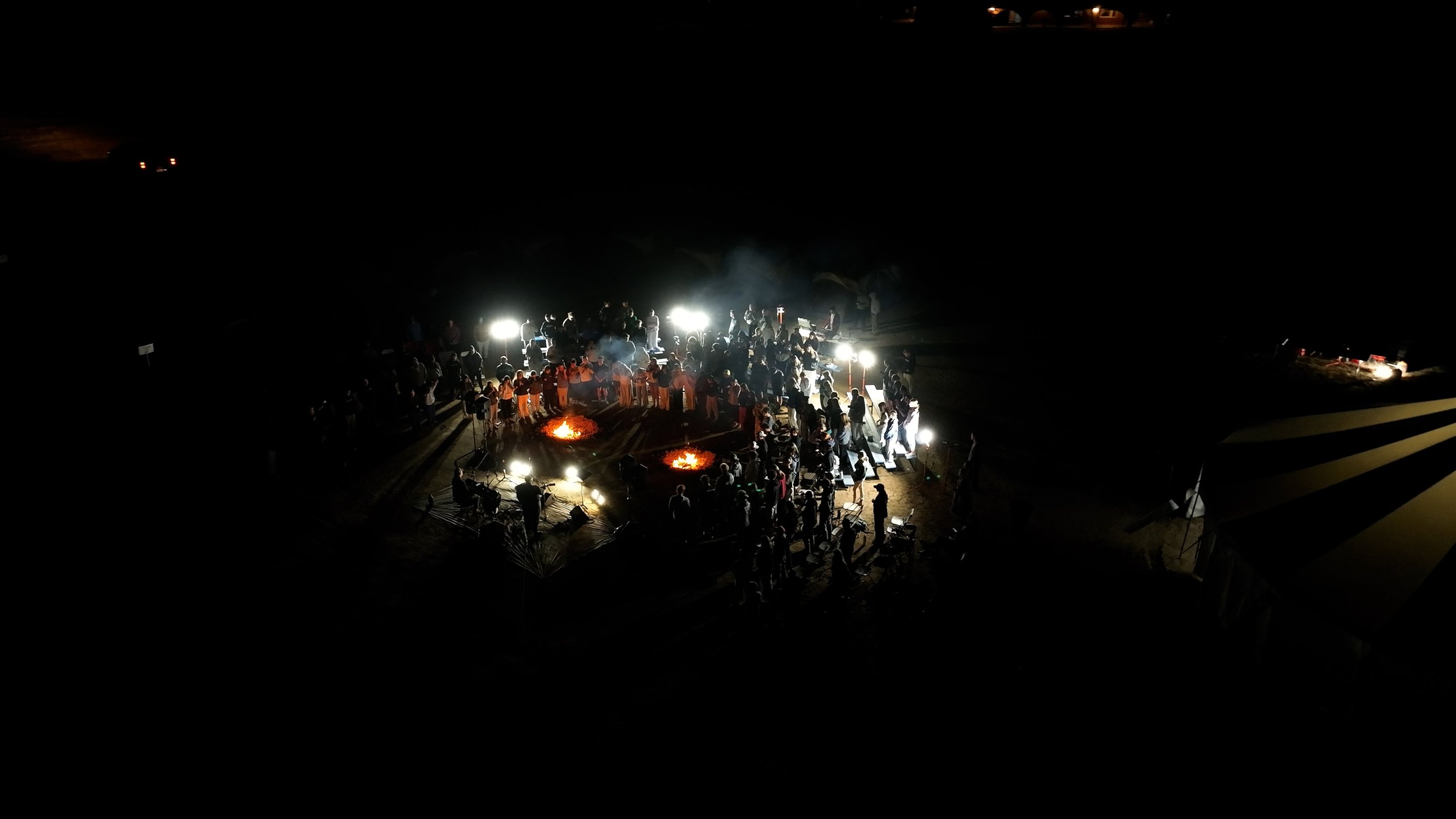 Night outdoor gathering with people around two fires, bright lights illuminating the scene, and dark surroundings.