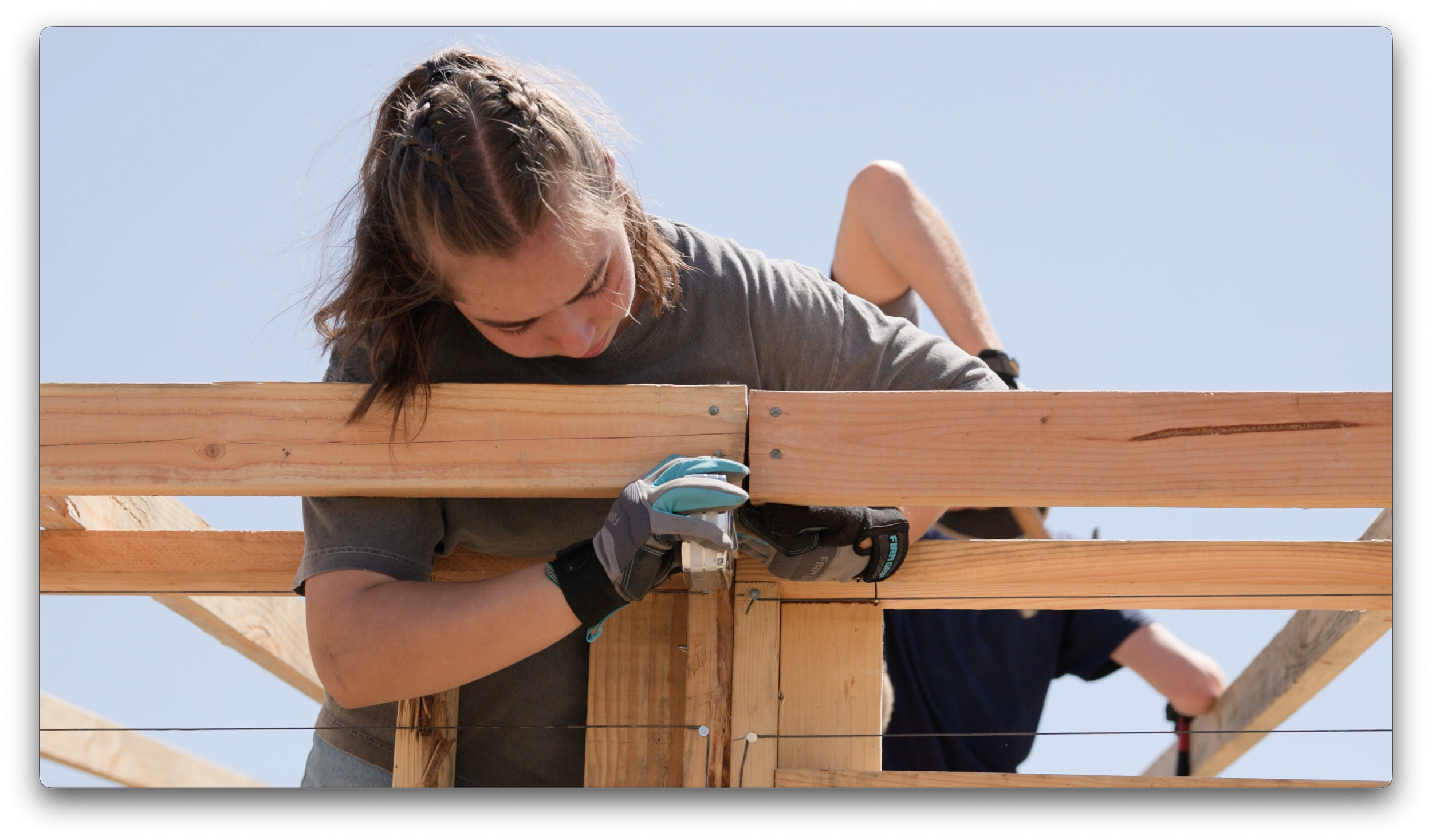 A woman working on a wooden construction project outdoors, using a power drill on a wooden beam, against a clear blue sky.