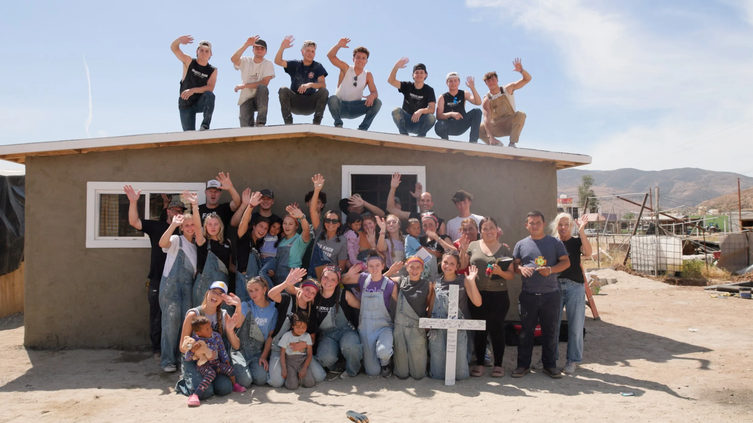 Group of people, including adults and children, gathered in front of a building under construction. Some are standing and waving, and some are sitting or kneeling. The building has a finished lower level and an unfinished upper level with people sitting on the roof. The background shows a rural landscape with mountains. The scene appears cheerful and celebratory.