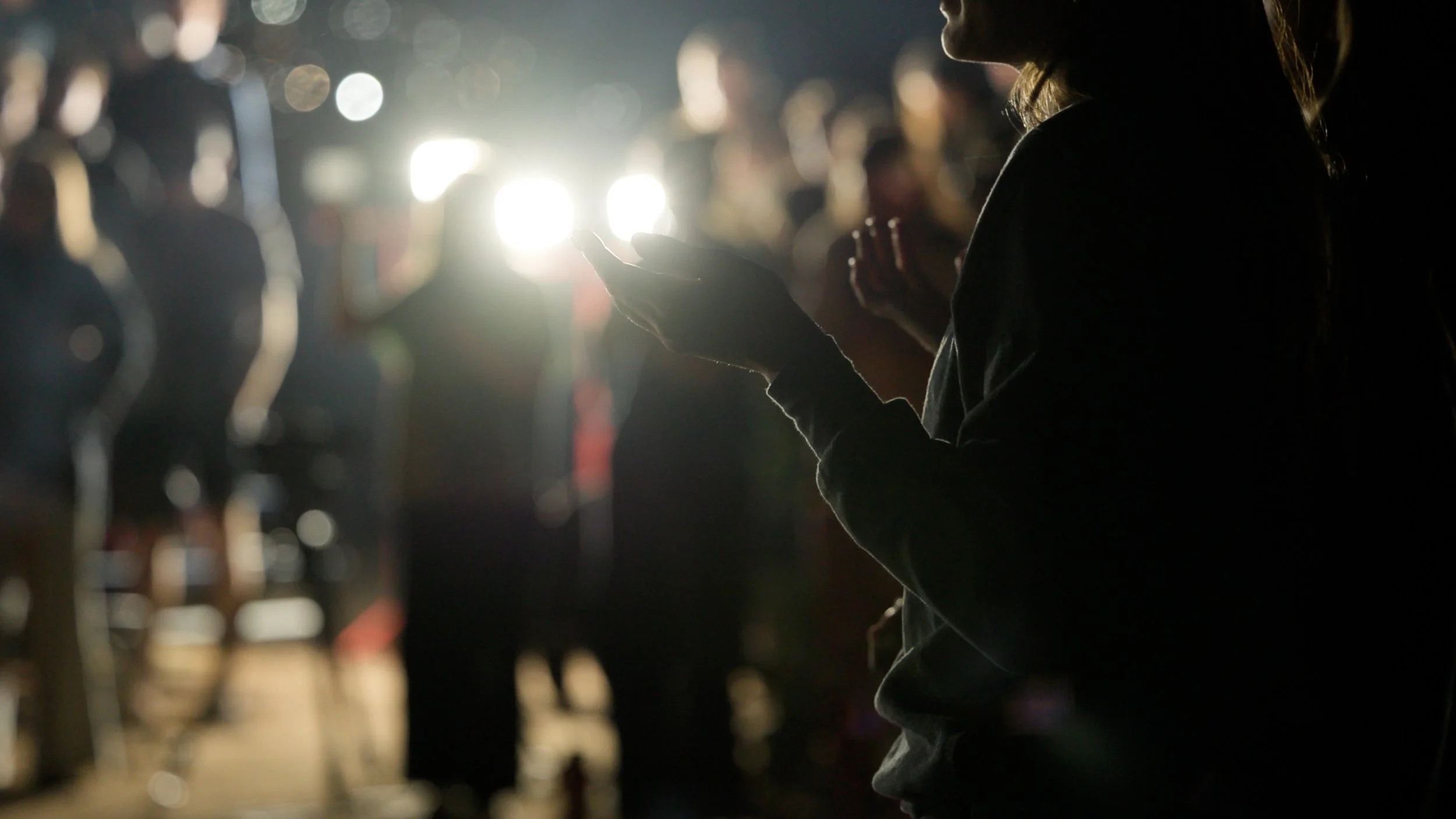 A person sitting and using a smartphone at night in a busy outdoor area with many blurred people in the background and bright lights behind.