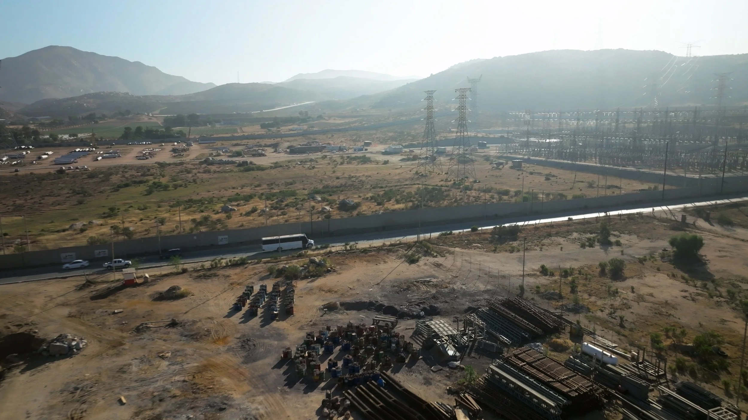 Scenic view of a hilly landscape with power lines, a highway, and construction site, under bright sunlight and hazy sky.