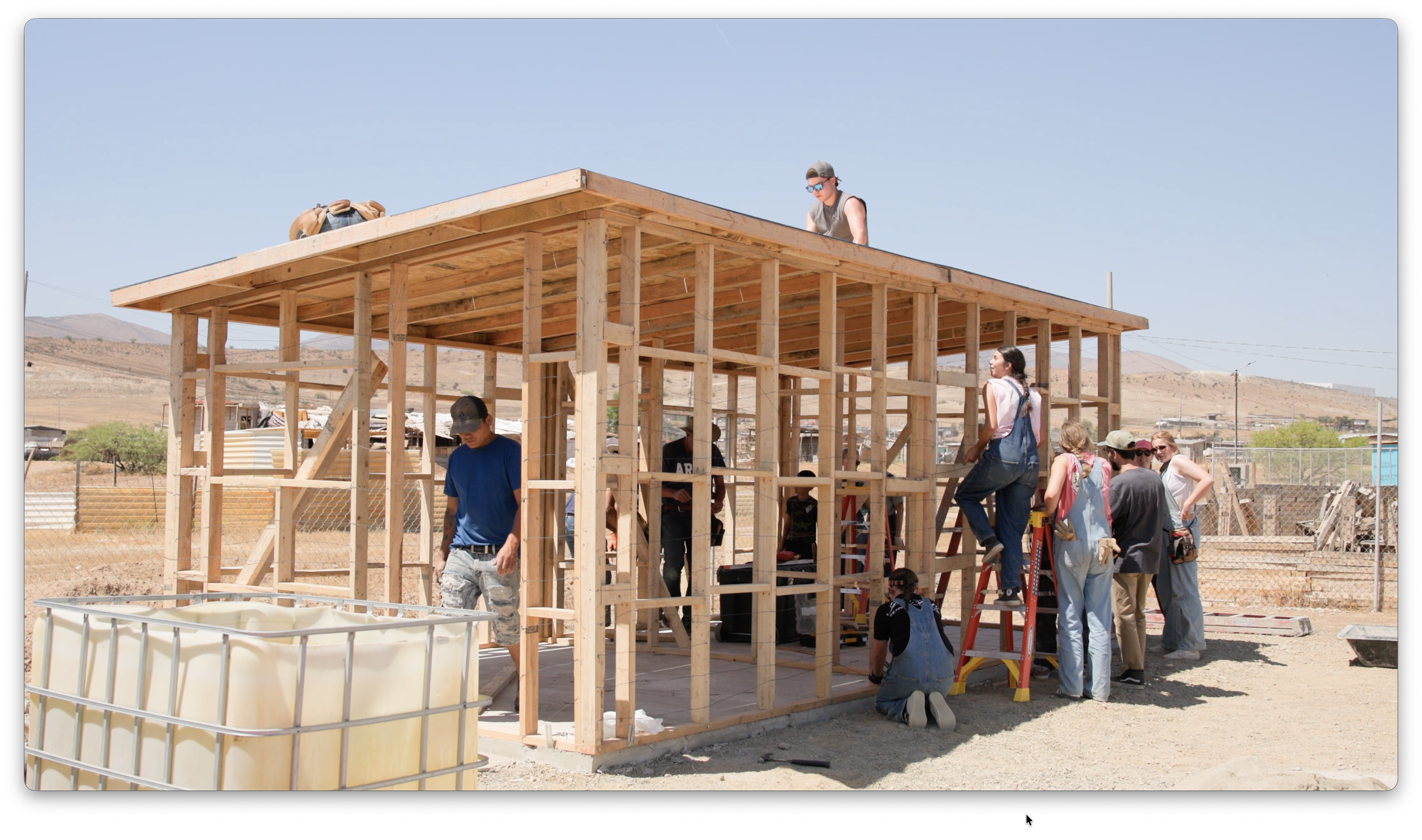 People building a wooden structure outdoors under a clear sky, with some using ladders and others observing.