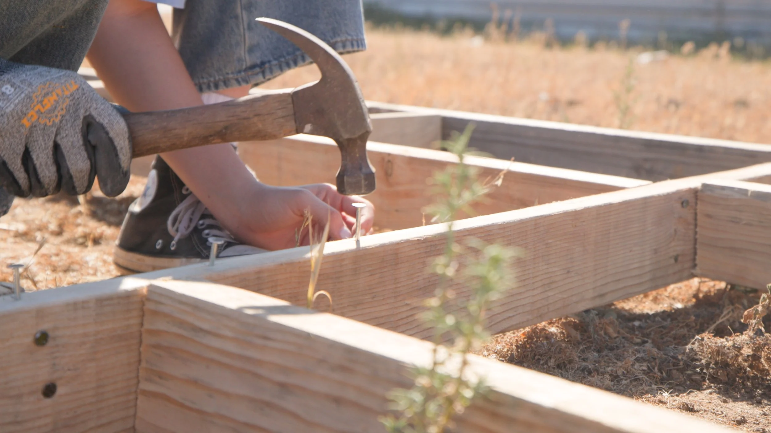 A person using a hammer to nail a nail into a wooden frame outdoors during daytime.