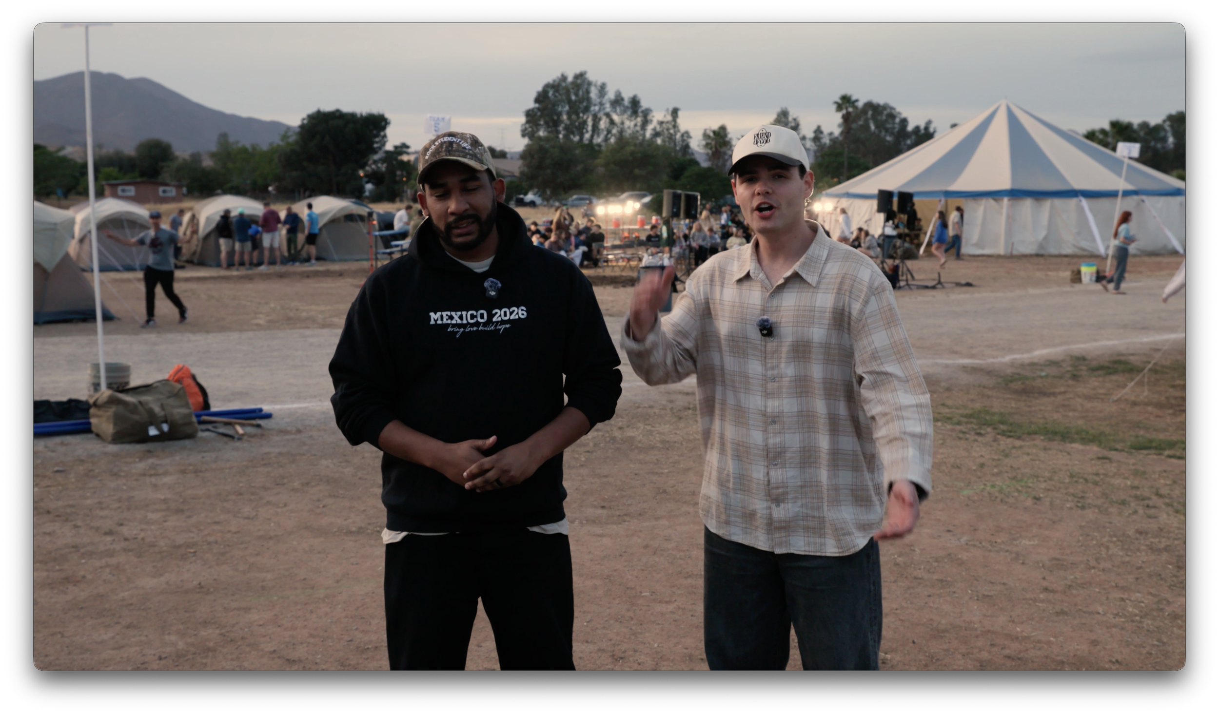 Two young men standing outdoors in front of a campsite, with tents and people in the background, during the evening. One is wearing a black hoodie with 'Mexico 2026' printed on it, and the other is in a beige plaid shirt and white cap, speaking to the camera.