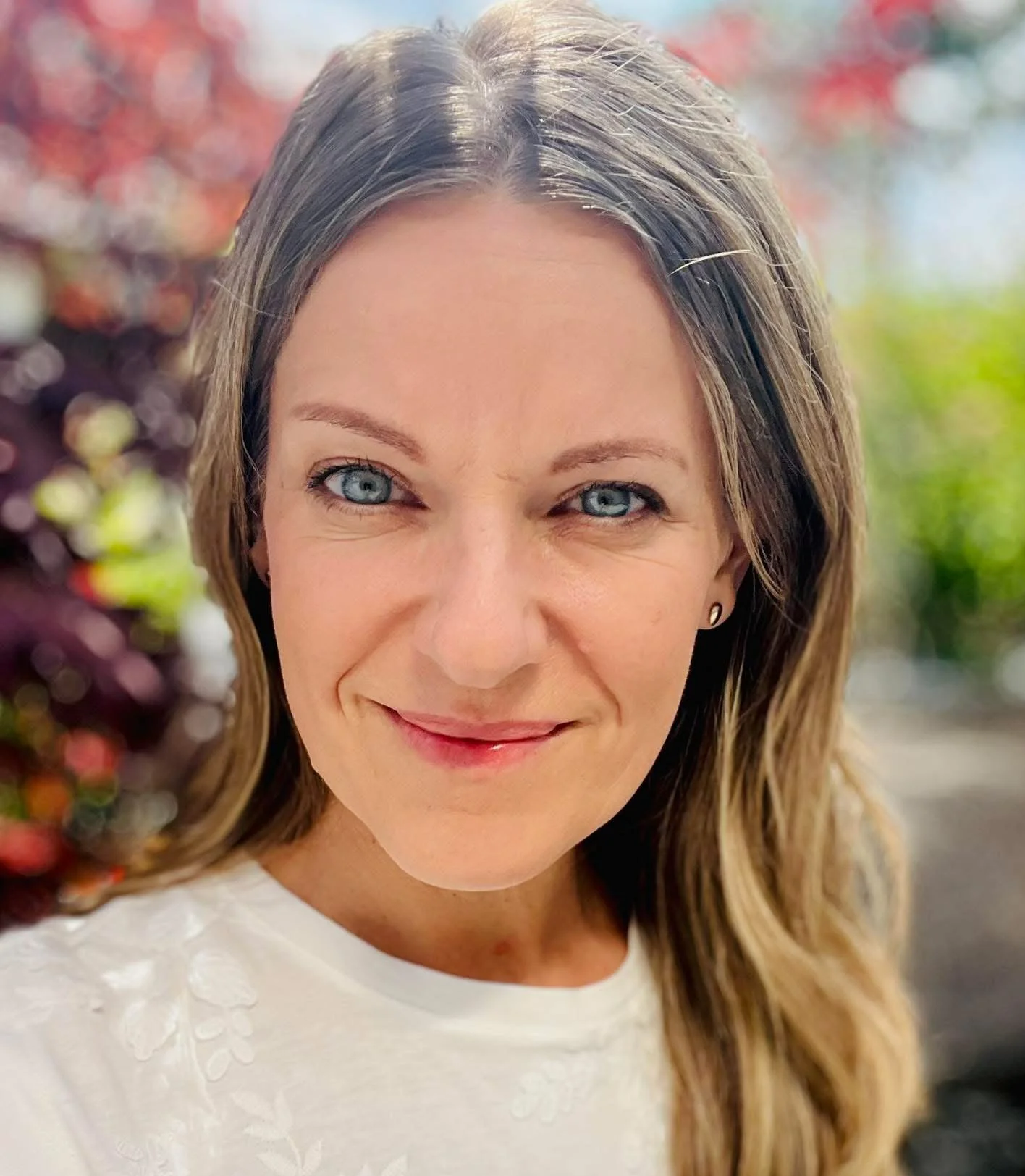 Nahaufnahme einer lächelnden Frau mit blauen Augen und braunen, gewellten Haaren vor einem bunten, unscharfen Hintergrund.