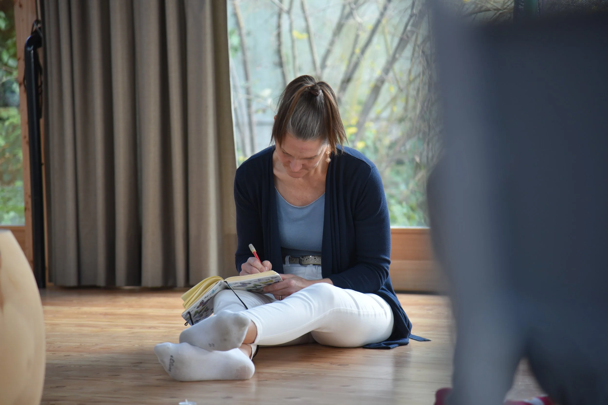 Ein Frau sitzt auf dem Boden vor einem großen Fenster mit Vorhängen und schreibt in ein Notizbuch.