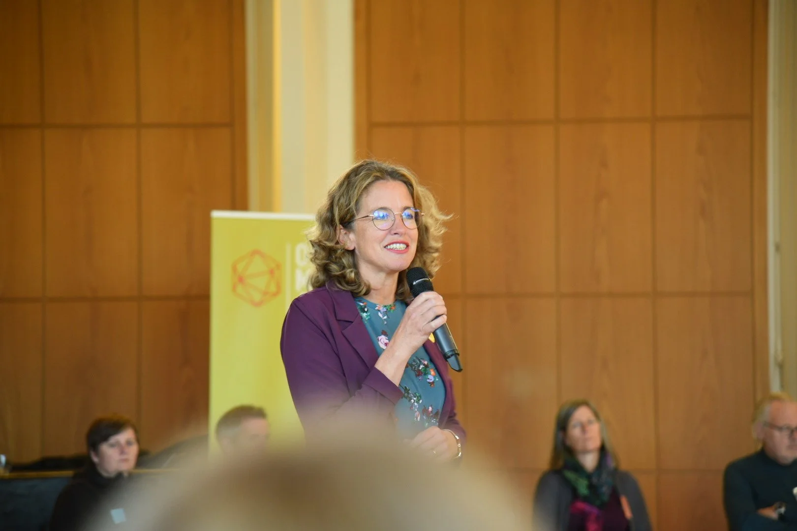 Eine Frau mit Brille hält ein Mikrofon und spricht bei einer Veranstaltung. Im Hintergrund ist eine gelbe Tafel mit einem roten geometrischen Symbol.