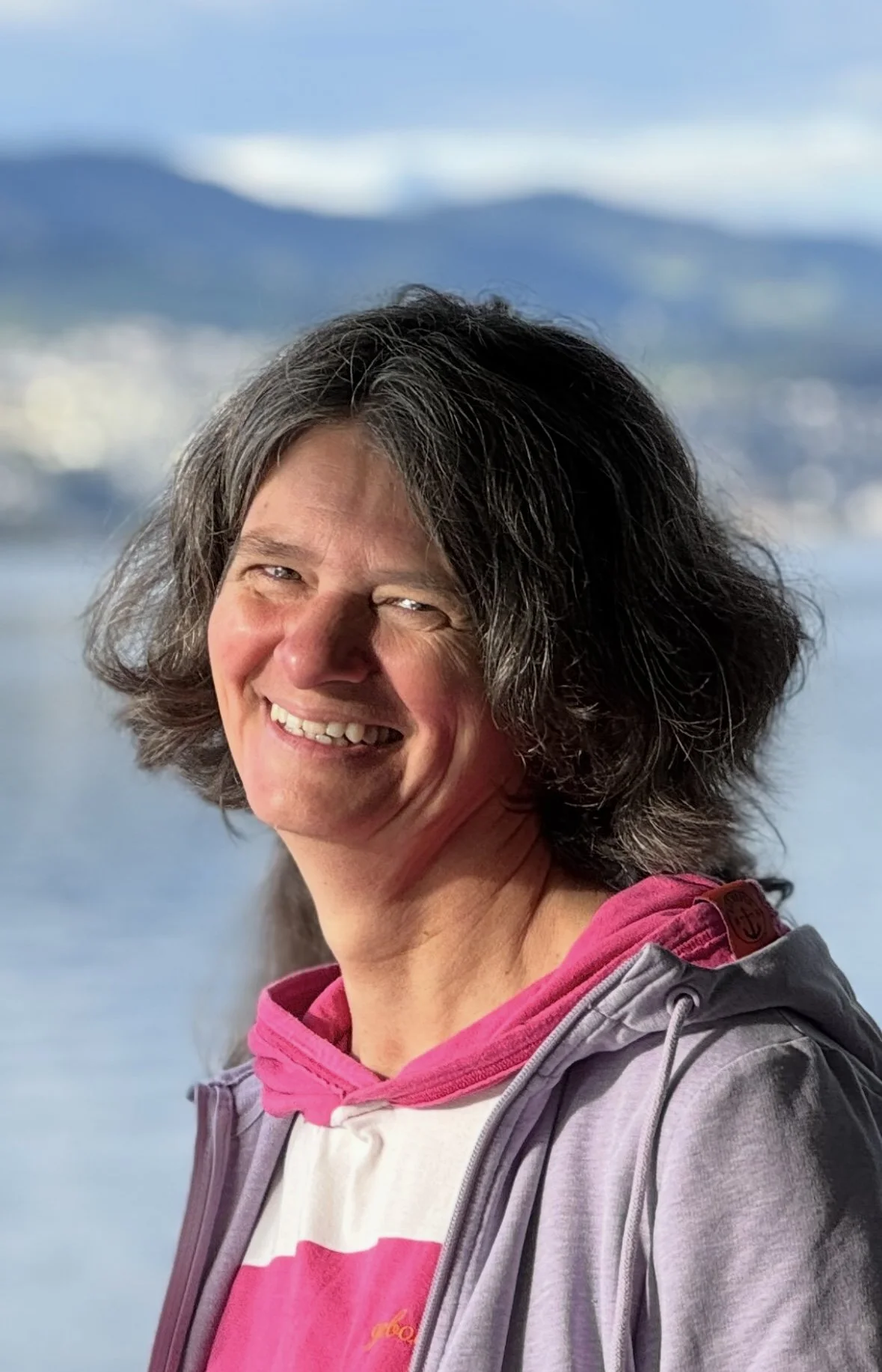 Lächelnde Frau mit kurzen, dunklen Haaren, trägt einen grauen Hoodie und eine pink-weiße Jacke, stehend vor einem Wasser- und Berglandschaft im Hintergrund.