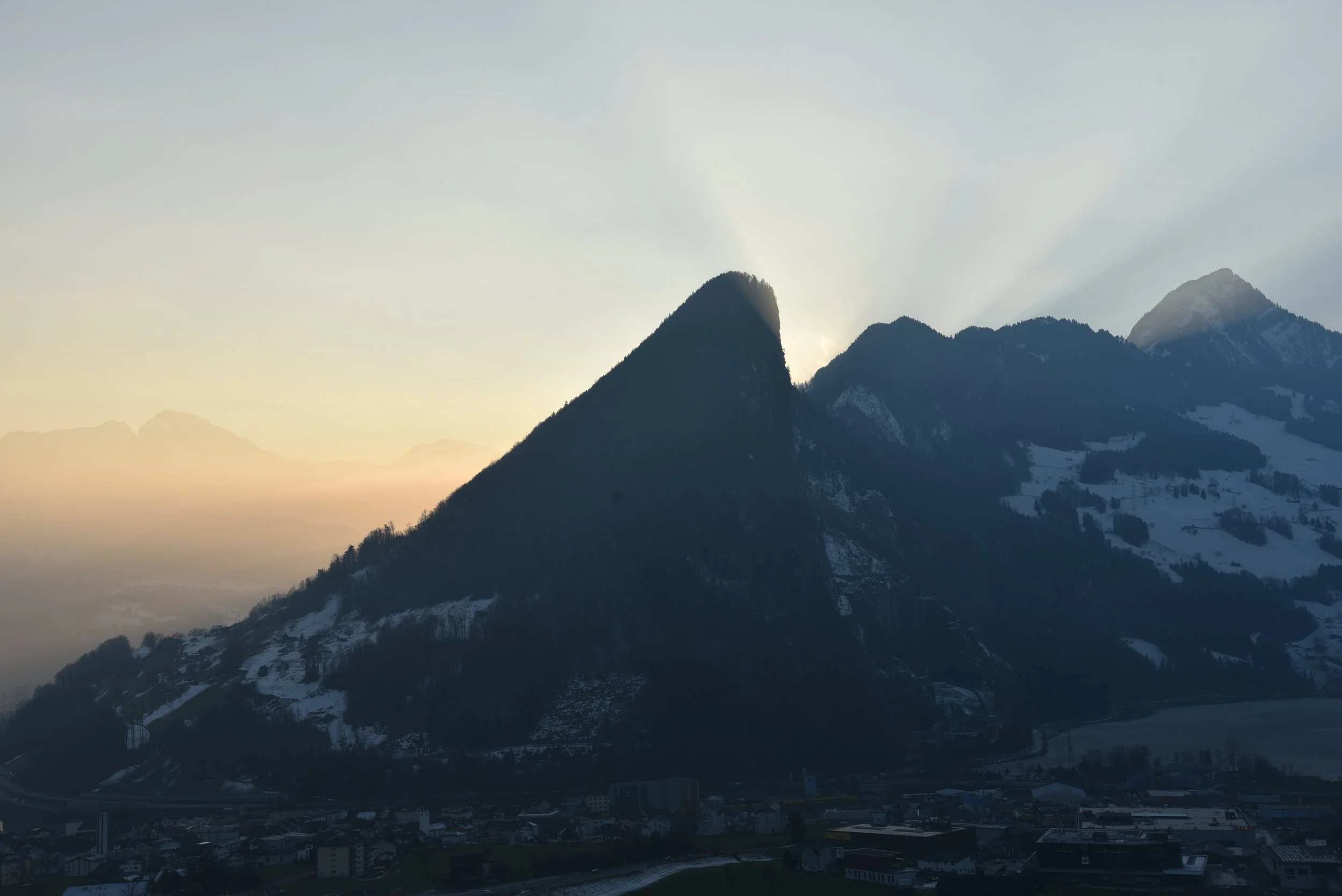 Berg im Sonnenuntergang mit dunkler Silhouette, Schneeflächen und einem kleinen Dorf im Vordergrund.