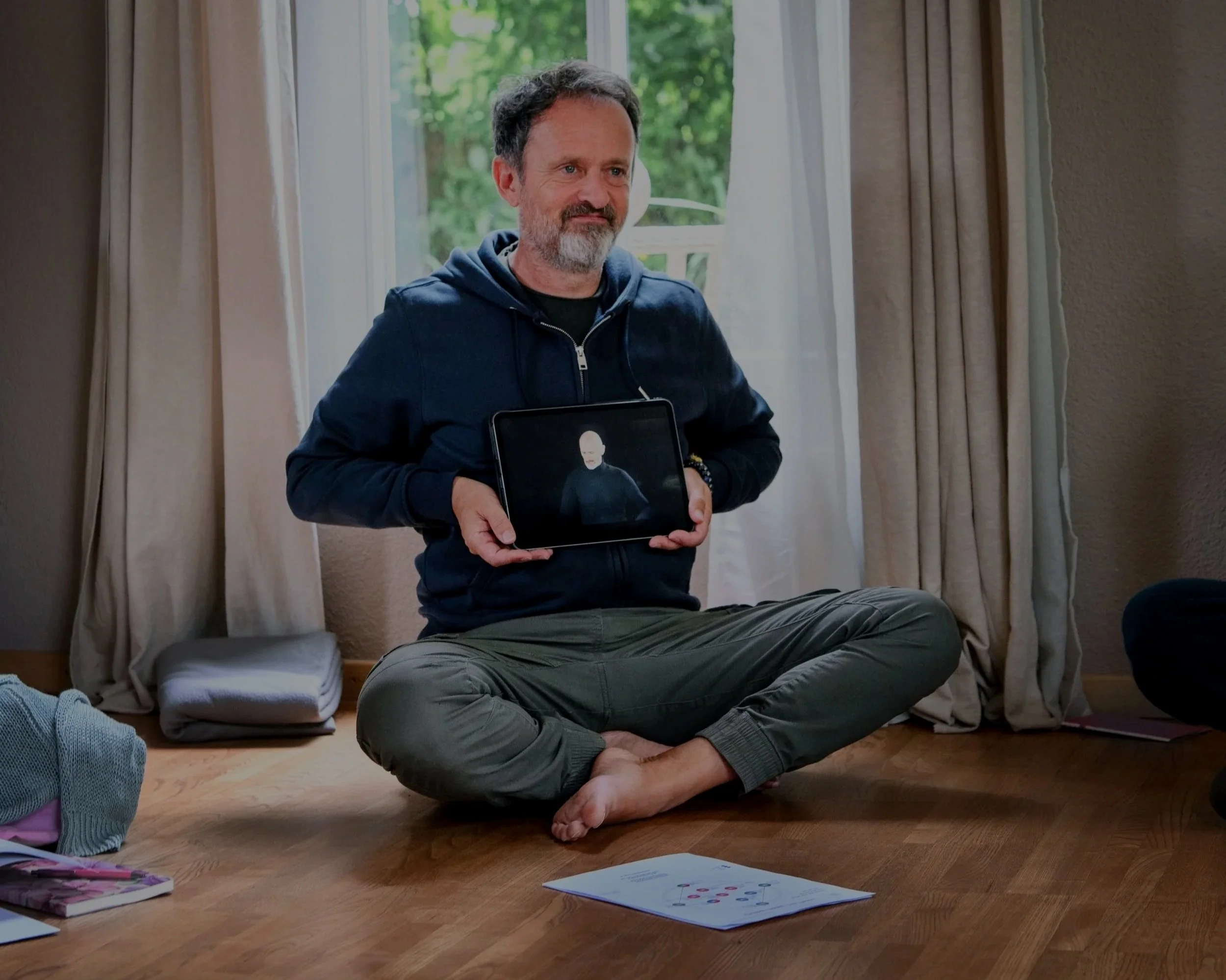 Ein Mann sitzt auf dem Holzboden in einem Wohnzimmer, hält ein Tablet mit einem Bild eines roten, kahlköpfigen Charakters und lächelt leicht. Im Vordergrund liegen Zeitschriften und eine Illustration.