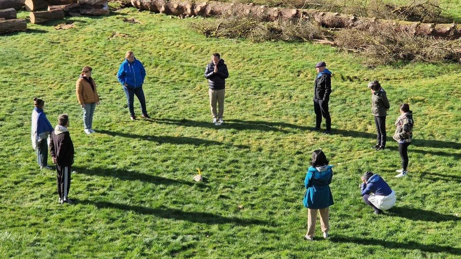 Gruppe von Menschen im Kreis auf einer grünen Wiese, einige beten, andere beobachten. Im Hintergrund sind Baumstämme und Sträucher. Es ist sonnig, Schatten sind sichtbar.