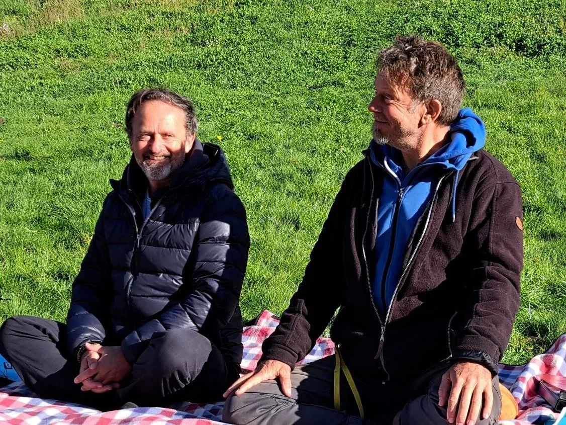 Zwei Männer sitzen auf einer Picknickdecke im Grünen, lachen und genießen das sonnige Wetter.