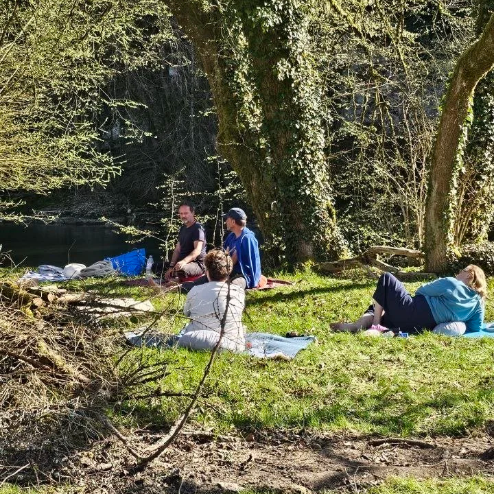 Menschen entspannen und sitzen auf einer Wiese im Wald neben einem Fluss, umgeben von Bäumen.