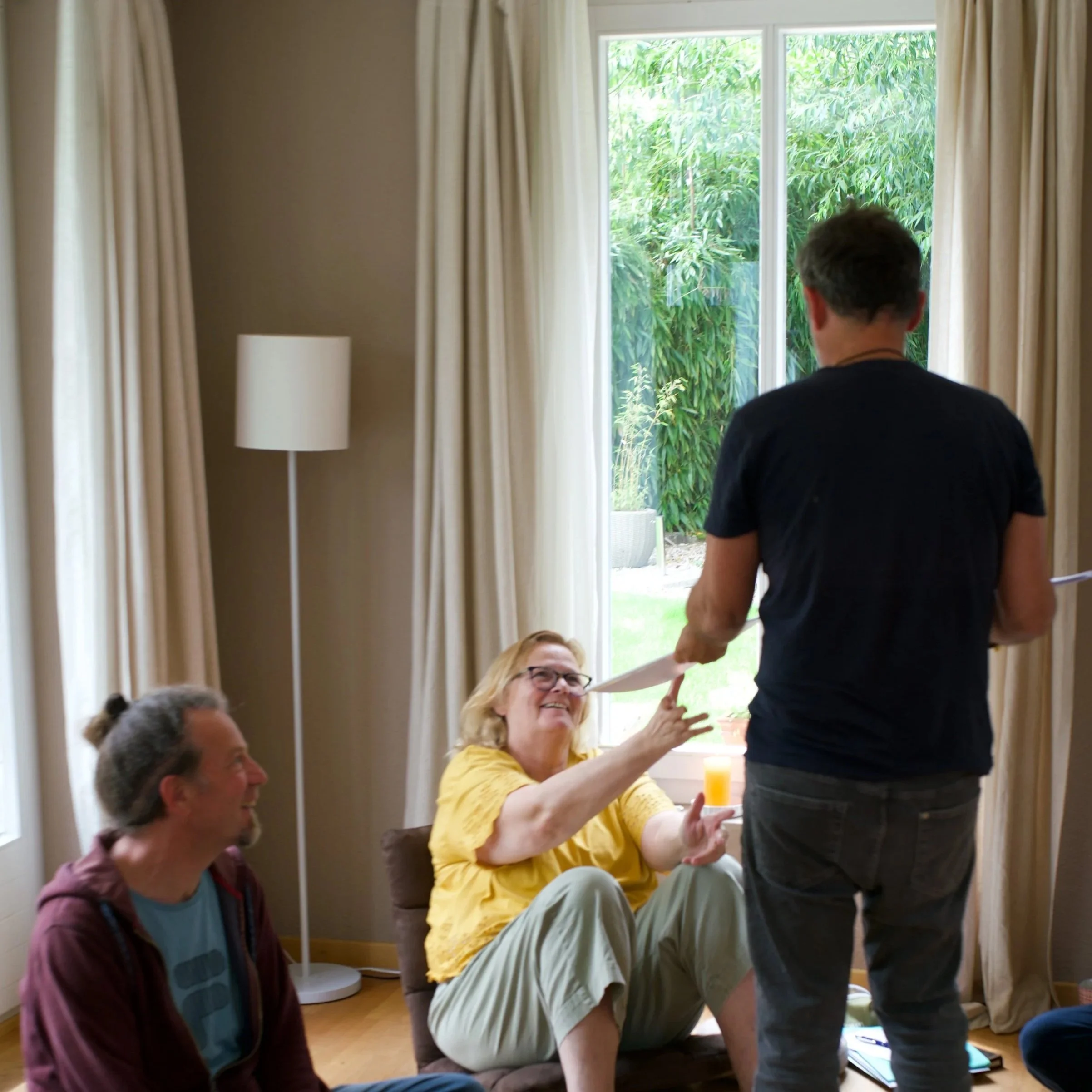 Drei Menschen sitzen im Wohnzimmer, eine Frau in gelbem Hemd reicht einem Mann einen Brief oder ein Dokument. Im Hintergrund ein großer Fenster mit Blick auf einen Garten, und eine stehende Lampe auf der linken Seite.