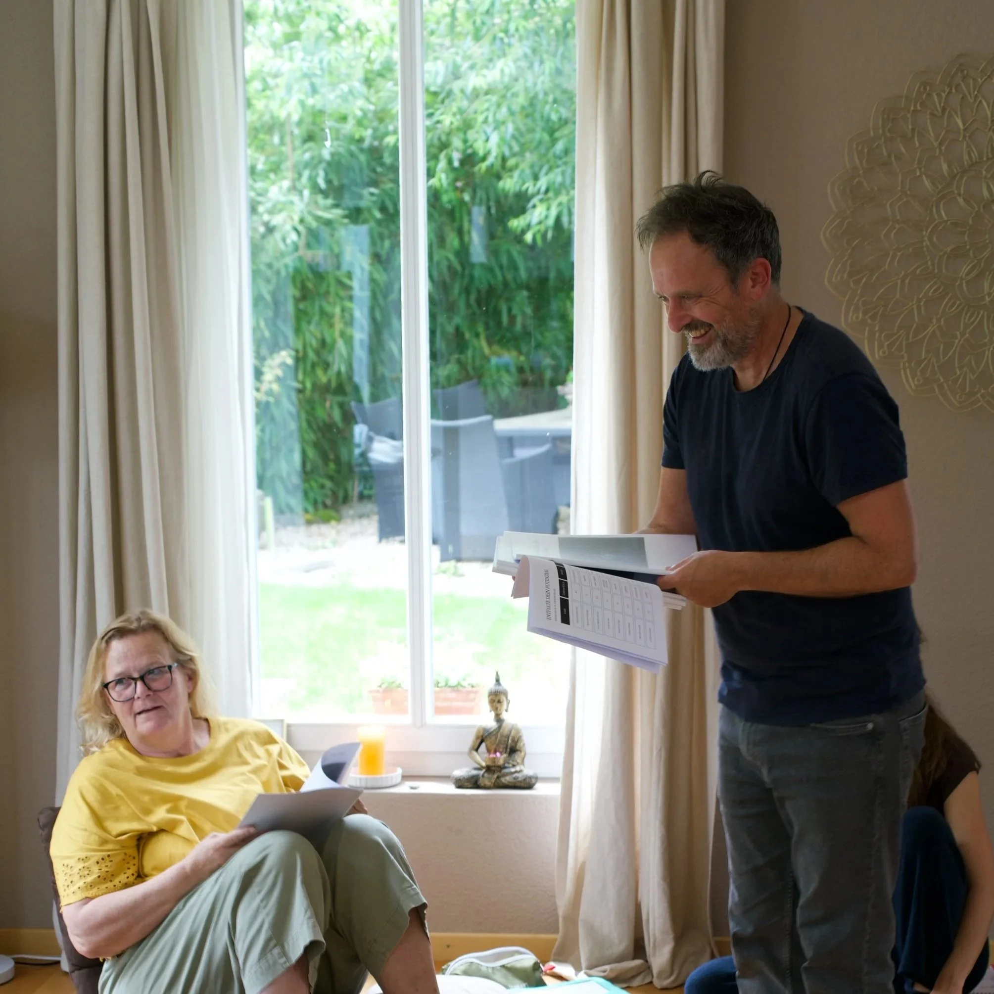 Ein Mann mit grauem Bart, dunklem T-Shirt und grauen Hosen steht lachend vor einer sitzenden Frau mit Gläsern und gelbem Hemd, während sie gemeinsam Dokumente prüfen. Im Hintergrund ein Fenster mit Blick auf einen Garten.