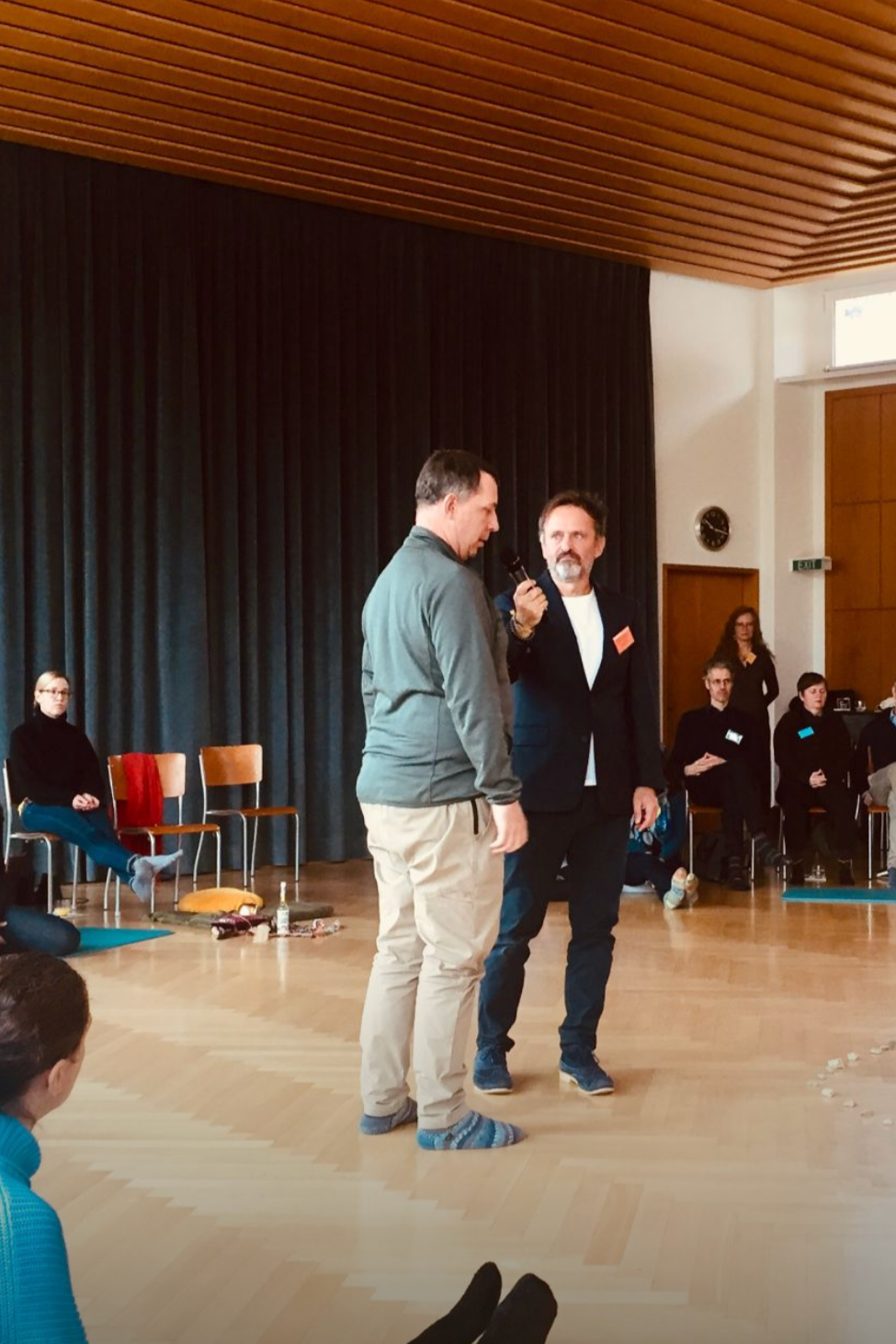 Zwei Männer stehen in einem Raum, einer hält ein Mikrofon, der andere wird interviewt oder spricht. Im Hintergrund sitzen weitere Personen, einige auf Stühlen, einige auf dem Boden. Der Raum hat dunkle Vorhänge, eine Holzdecke und einen hohen Raum.