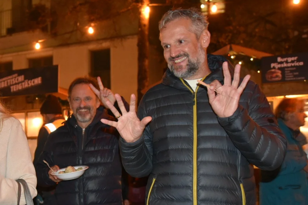 Zwei Männer in dunklen Jacken bei einer Veranstaltung im Freien, einer macht eine Handsignale und die andere hält eine Schüssel mit Essen, im Hintergrund sind Lichter und ein Food-Stand.