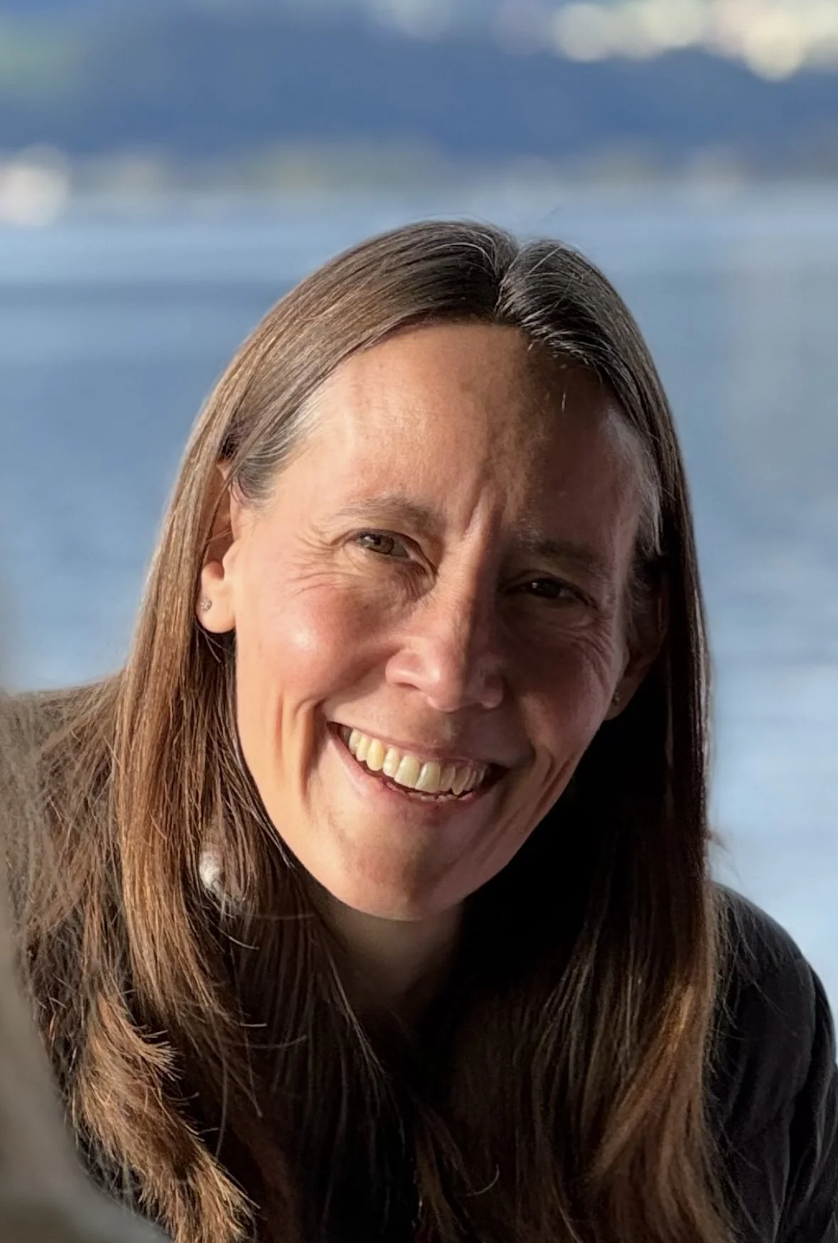 Lächelnde Frau mit braunen Haaren vor einem Wasserhintergrund.