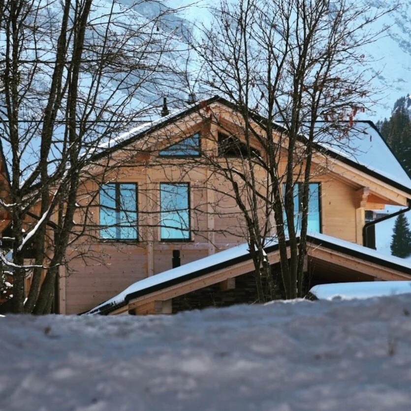 Holzhaus im Winter mit Schneedecke, umgeben von Bäumen ohne Blätter, blauer Himmel im Hintergrund.