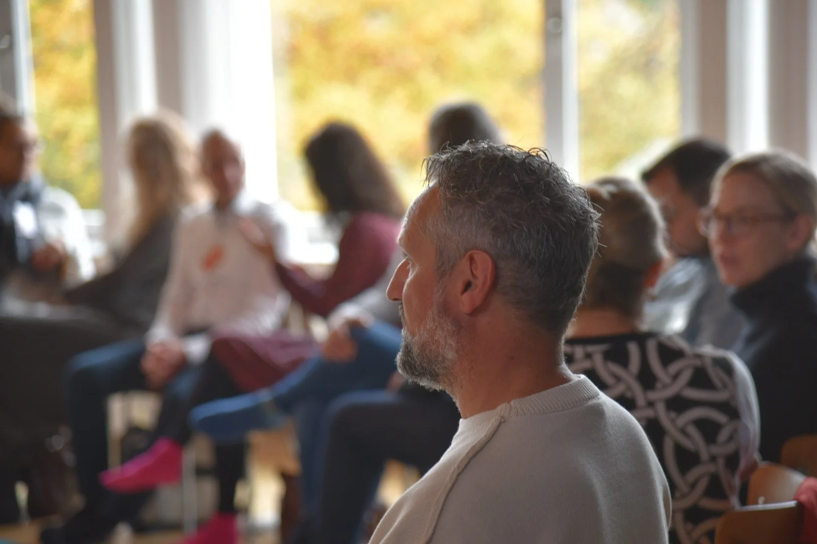 Menschen bei einer Diskussion oder einem Treffen in einem hell erleuchteten Raum mit großen Fenstern, im Hintergrund sind herbstliche Bäume zu sehen.