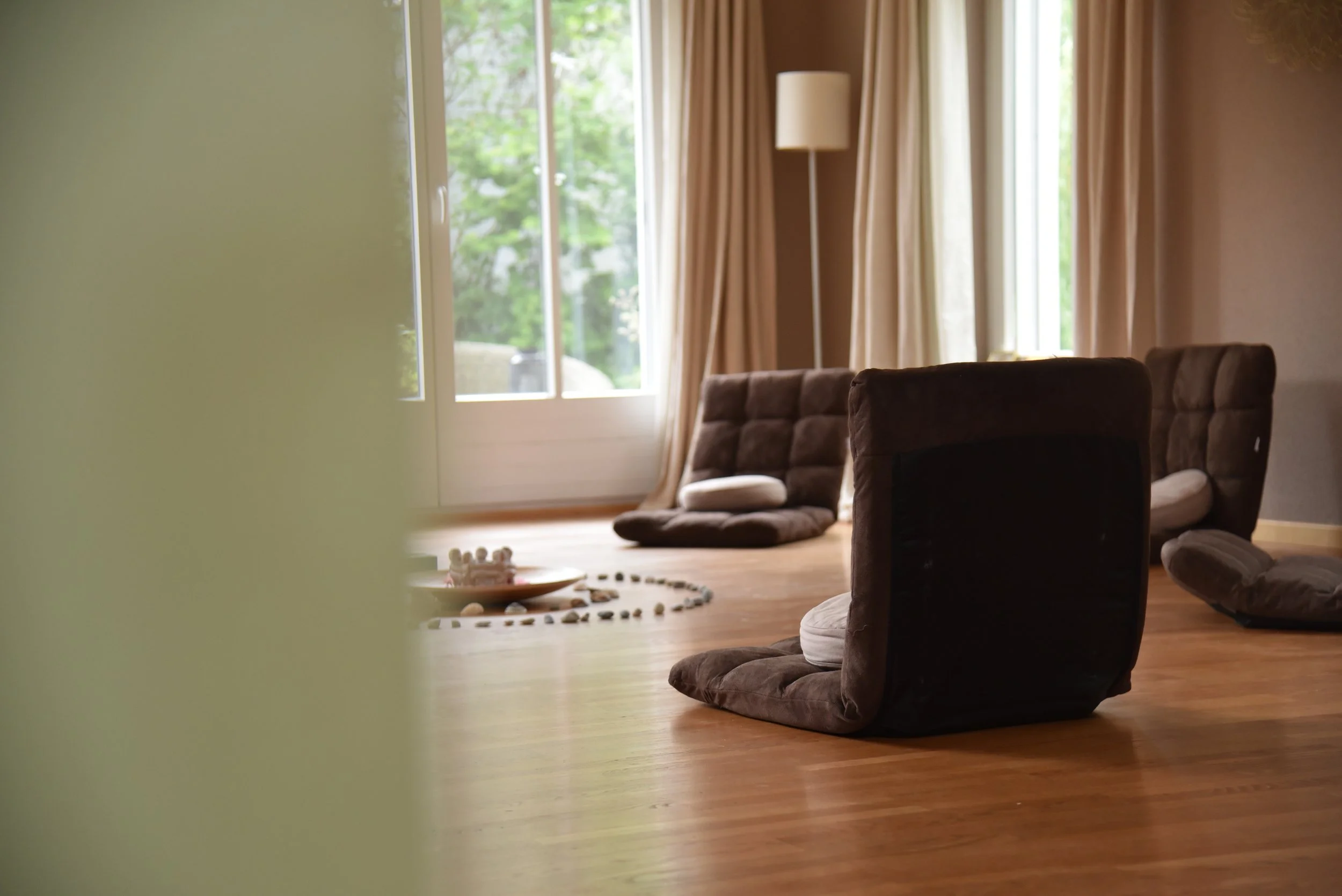 Ein Wohnzimmer mit Bodenkissen, einem runden Spielzeug, das auf dem Boden liegt, und einem großen Fenster mit Blick nach draußen, umgeben von Vorhängen.